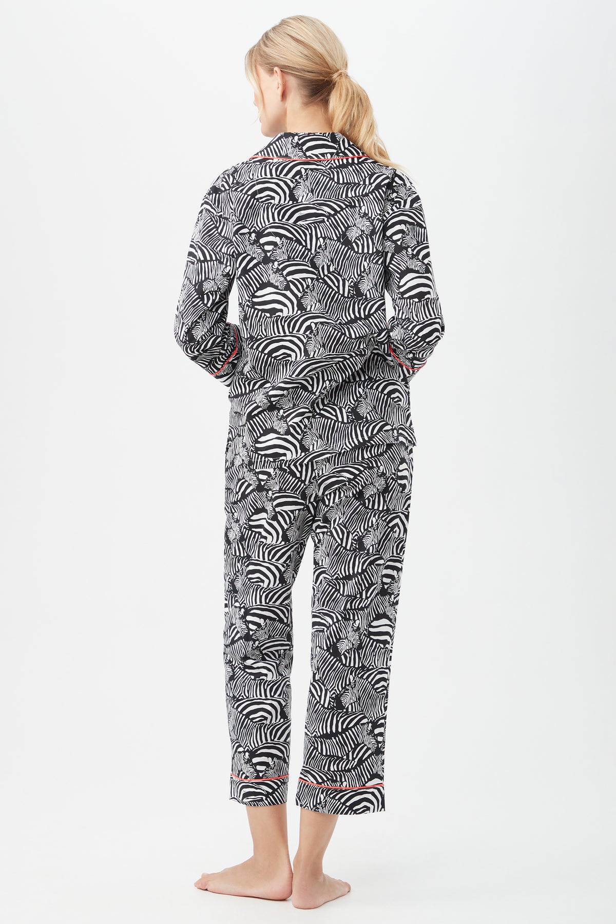 A blonde woman stands barefoot against a white background, modeling the Zebra Safari Womens 3/4-Sleeve Cropped Pant Poplin PJ Set—featuring black-and-white zebra stripes with red trim—perfect for chic womens sleepwear.