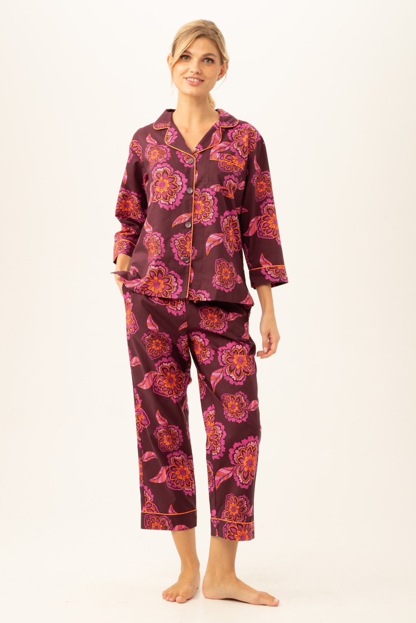 A woman smiles barefoot in the African Floral Women’s 3/4-Sleeve Cropped Pant Poplin PJ Set, featuring a vibrant multi-color floral print, a button-down top with 3/4 sleeves, and relaxed cropped pants with pockets—stylish and comfortable.