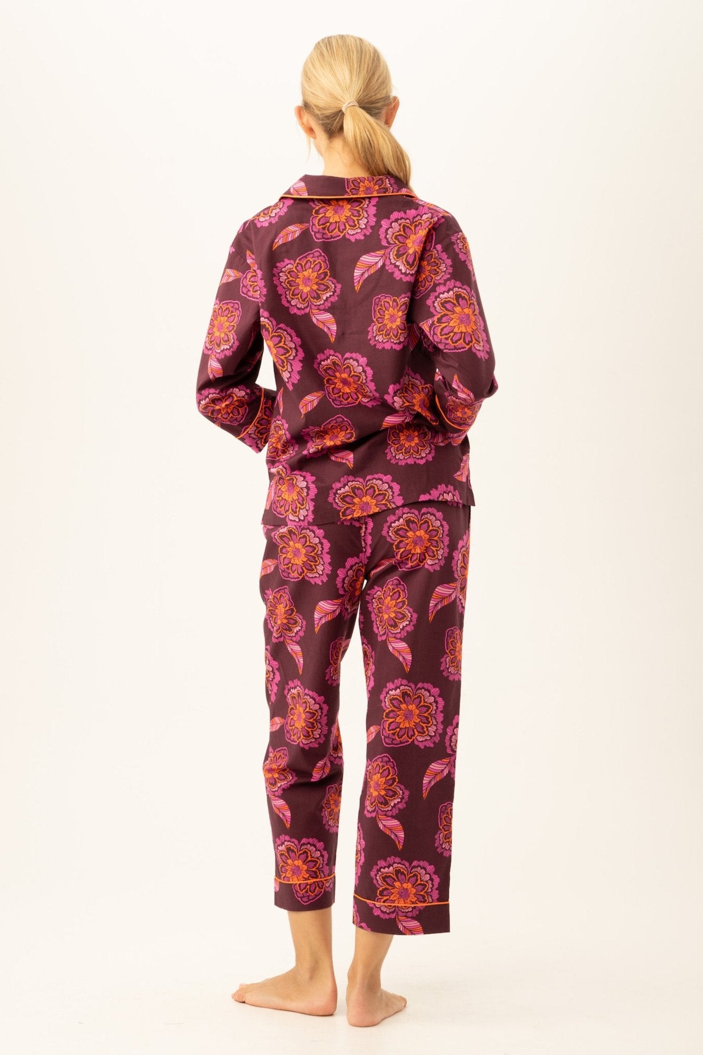 A woman stands barefoot facing away, wearing the African Floral Womens 3/4-Sleeve Cropped Pant Poplin PJ Set featuring a bold pink, orange, and purple floral print on a dark background. Her blonde hair is styled in a low ponytail.