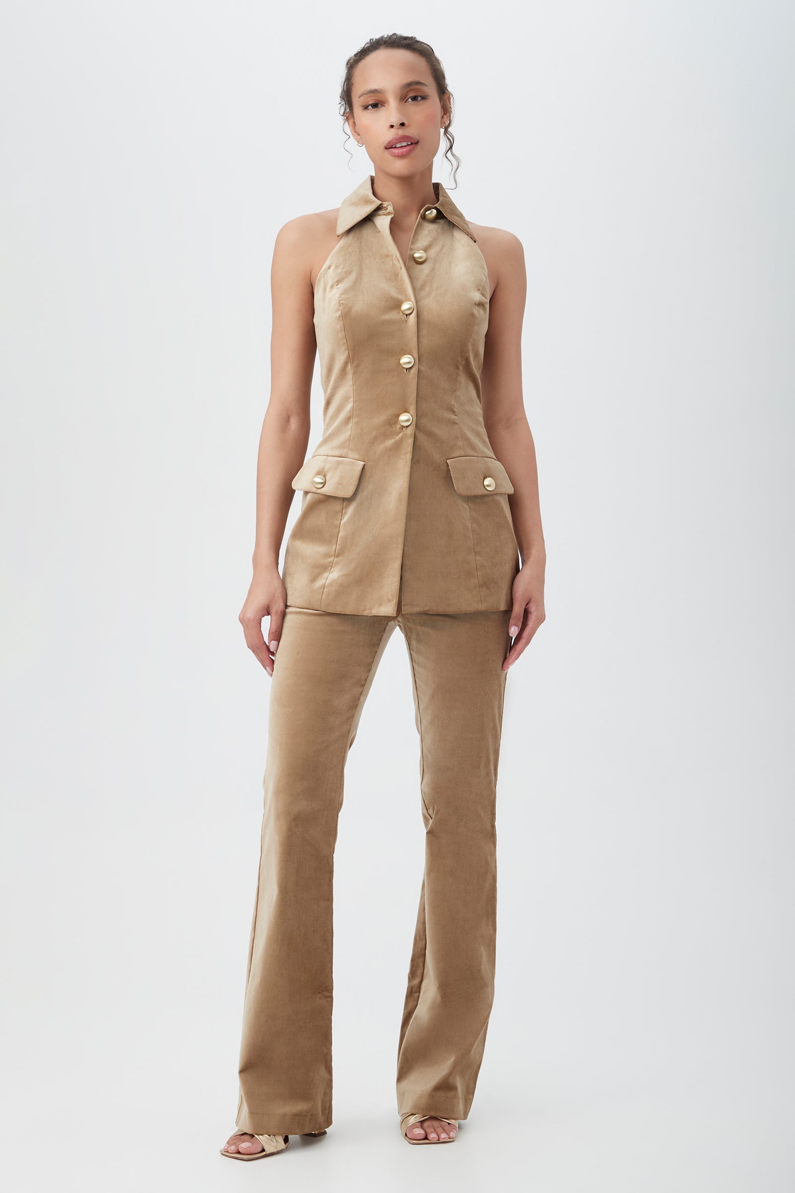 A woman stands against a white background wearing the Elainia Velveteen Sleeveless Tunic Top in tan with gold buttons and matching flared pants. Her hair is pulled back, and she pairs the outfit with open-toed heels.