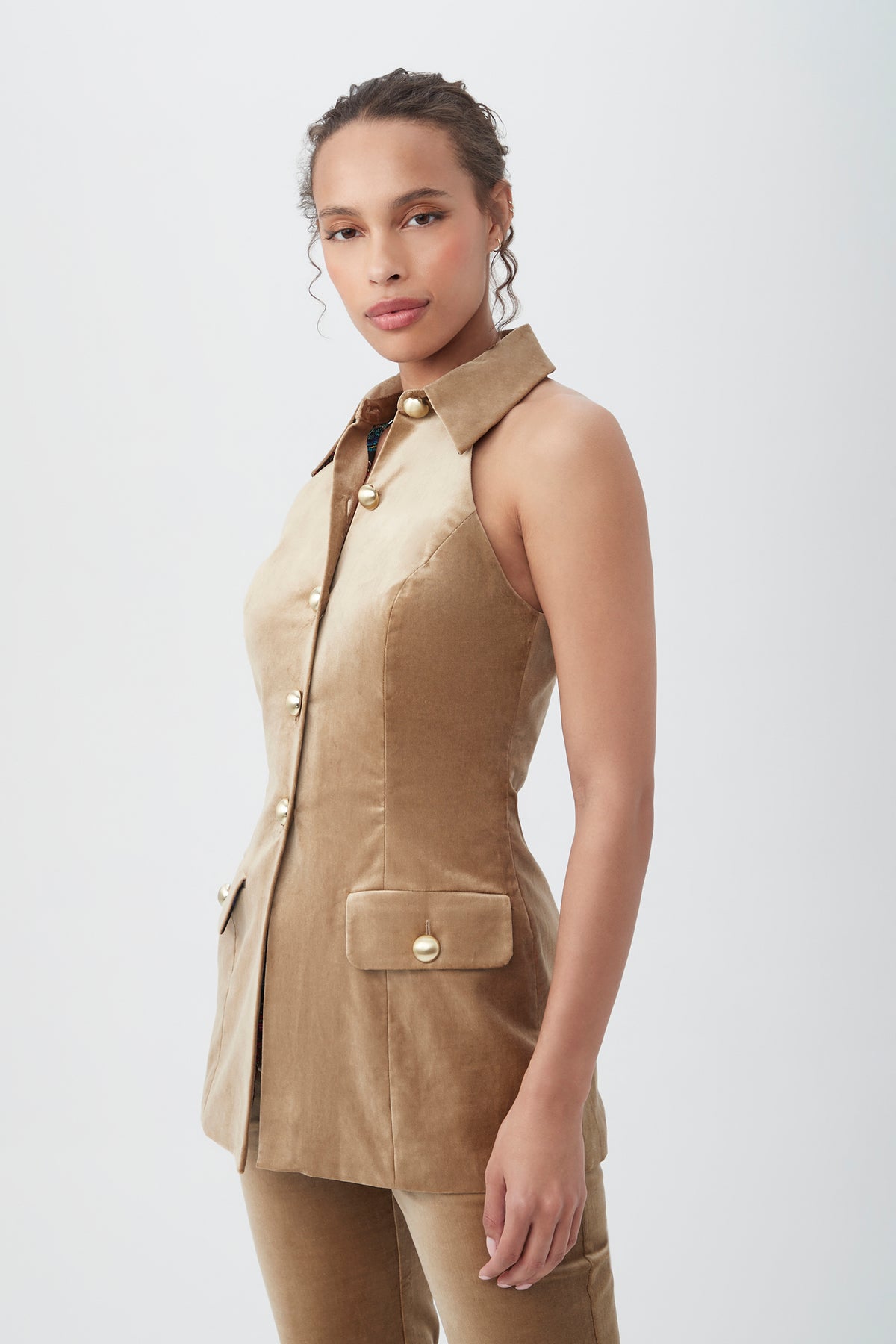 A woman with curly hair pulled back wears the Elainia Velveteen Sleeveless Tunic Top in gold with large buttons and flap pockets, paired with matching pants, standing against a plain white background.