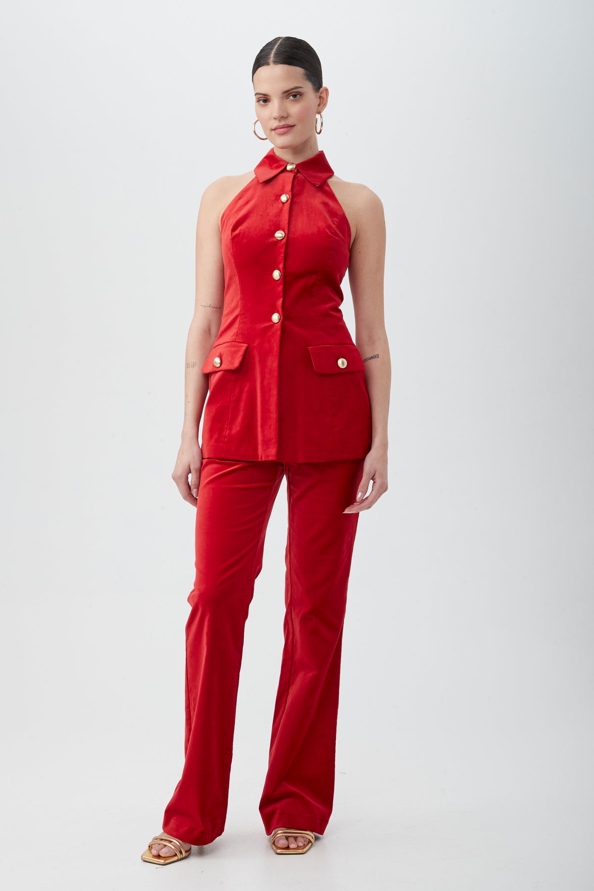 A woman stands against a white background wearing the Elainia Velveteen Sleeveless Tunic Top in red with a collar and matching pants. Her hair is tied back, and she accessorizes with hoop earrings and open-toe sandals.