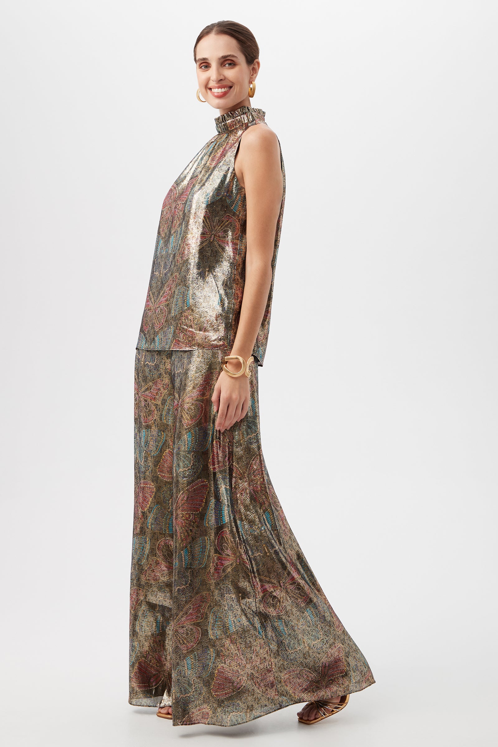 A woman smiles in the Rissa Butterfly Print Silk Sleeveless Mockneck Top with matching wide-leg pants, gold hoop earrings, a bracelet, and sandals against a white background.