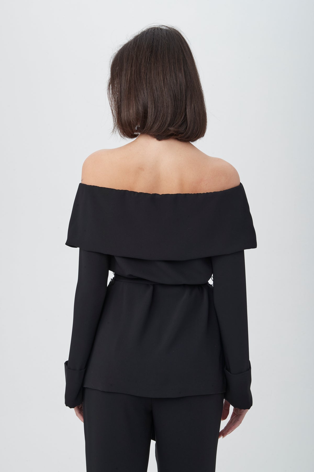 A woman with shoulder-length brown hair, seen from the back, wears the Lyndi Off-The-Shoulder Belted Long Sleeve Top and matching black pants against a plain white background.