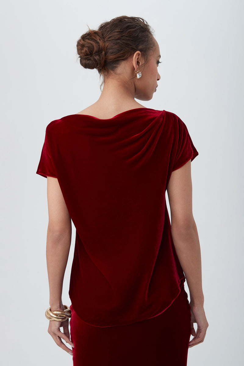 A woman with her hair in a bun wears the Sintha Velvet Boatneck Short Sleeve Top in deep red. She is facing away, revealing the draped back. She accessorizes with gold earrings and bracelets against a plain light background.