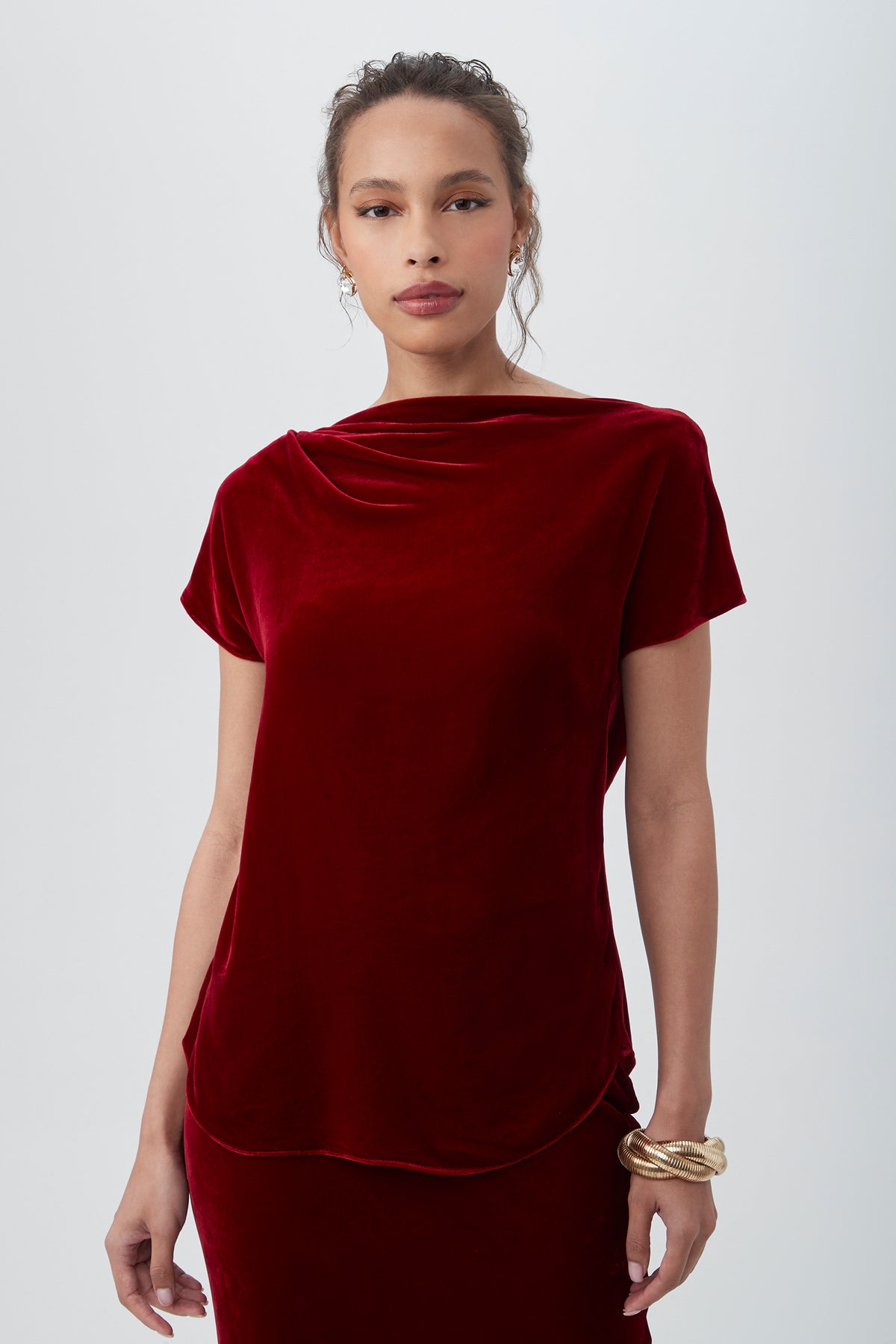 A woman with curly hair pulled back models the Sintha Velvet Boatneck Short Sleeve Top in Vixen Red against a light gray background, accessorized with gold bangles on her left wrist.