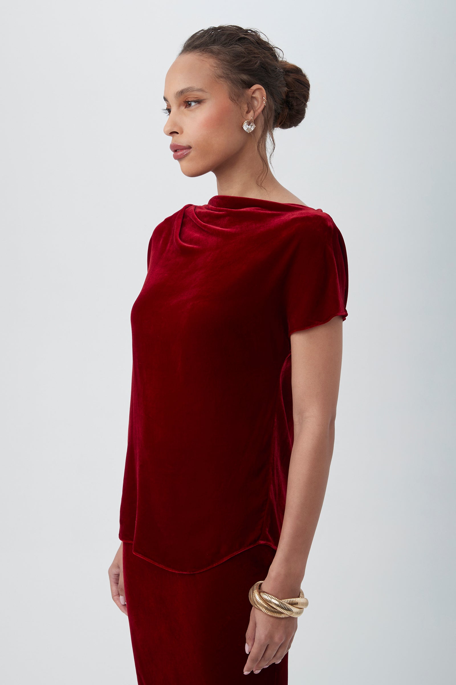 A woman stands in profile against a light background, wearing the Sintha Velvet Boatneck Short Sleeve Top in red, gold earrings and bangles, her hair styled neatly in a bun.