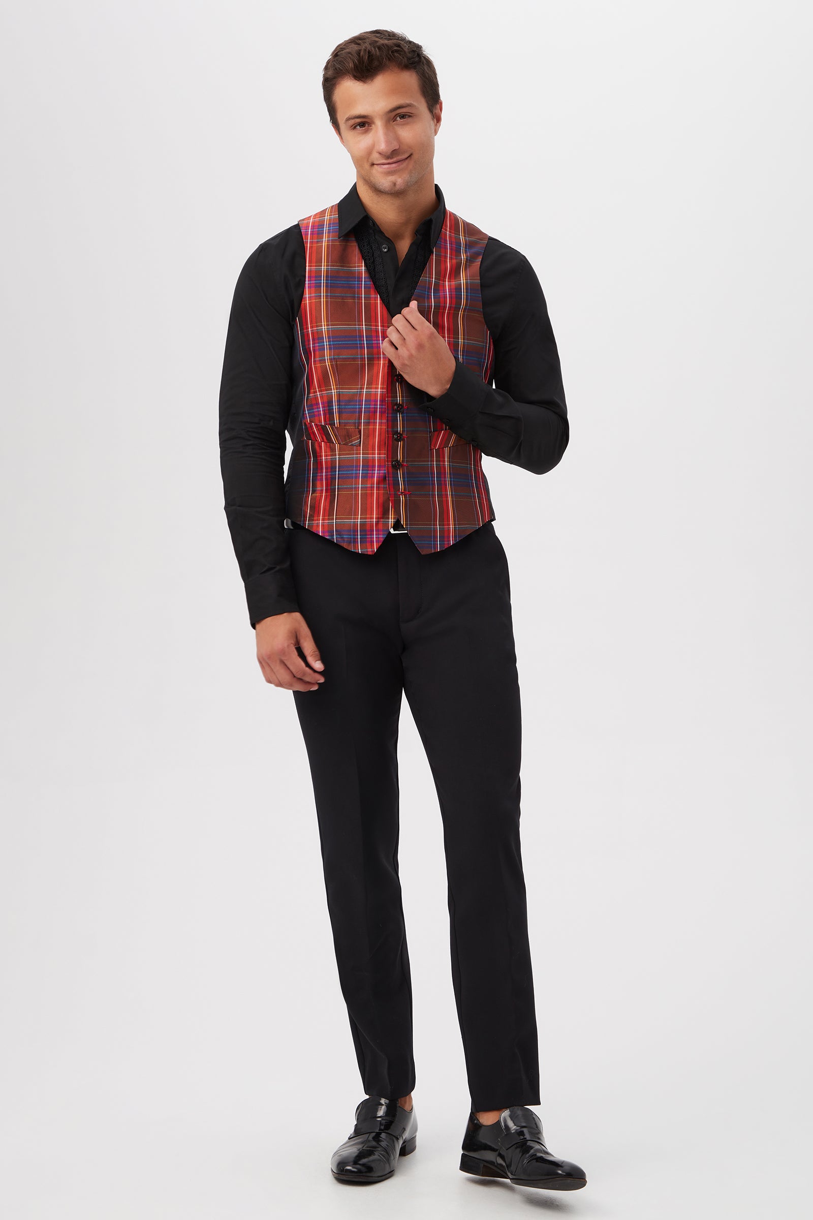 A man stands against a plain background wearing black Alex Slim Trouser - Luxe Drape, a black shirt, black dress shoes, and a red and orange plaid vest. He holds the lapel of his vest and smiles slightly.
