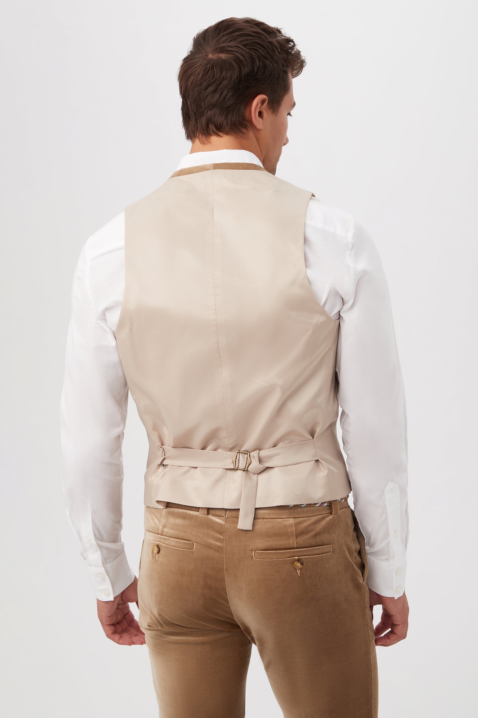 A man is shown from the back wearing the Irving Vest - Gold Velveteen over a white dress shirt and light brown trousers. The vest features an adjustable waist strap. The background is plain white.