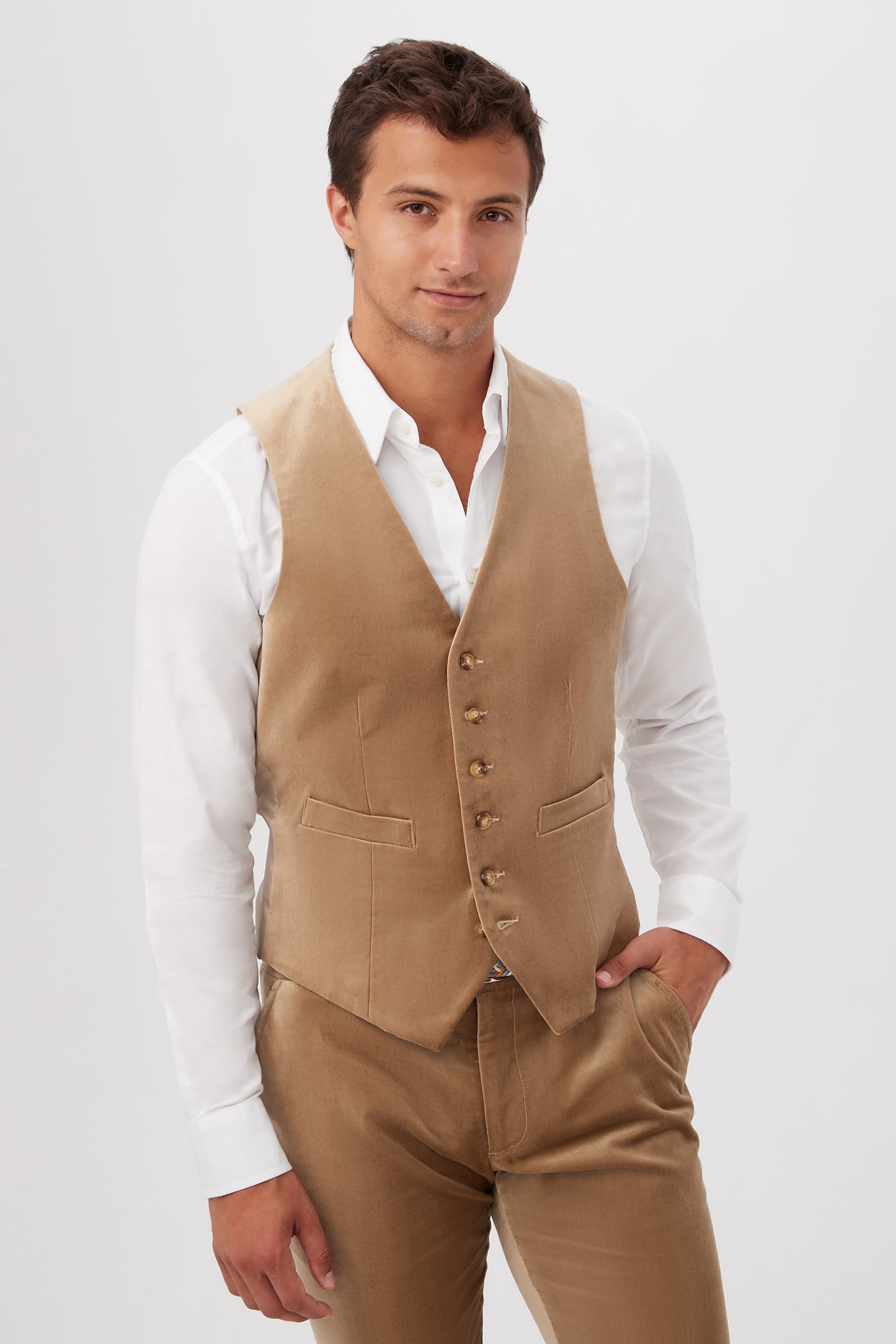 A young man with short brown hair stands against a plain background, smiling slightly with one hand in his pocket. He wears a white dress shirt, the Irving Vest in gold velveteen, and matching tan trousers.