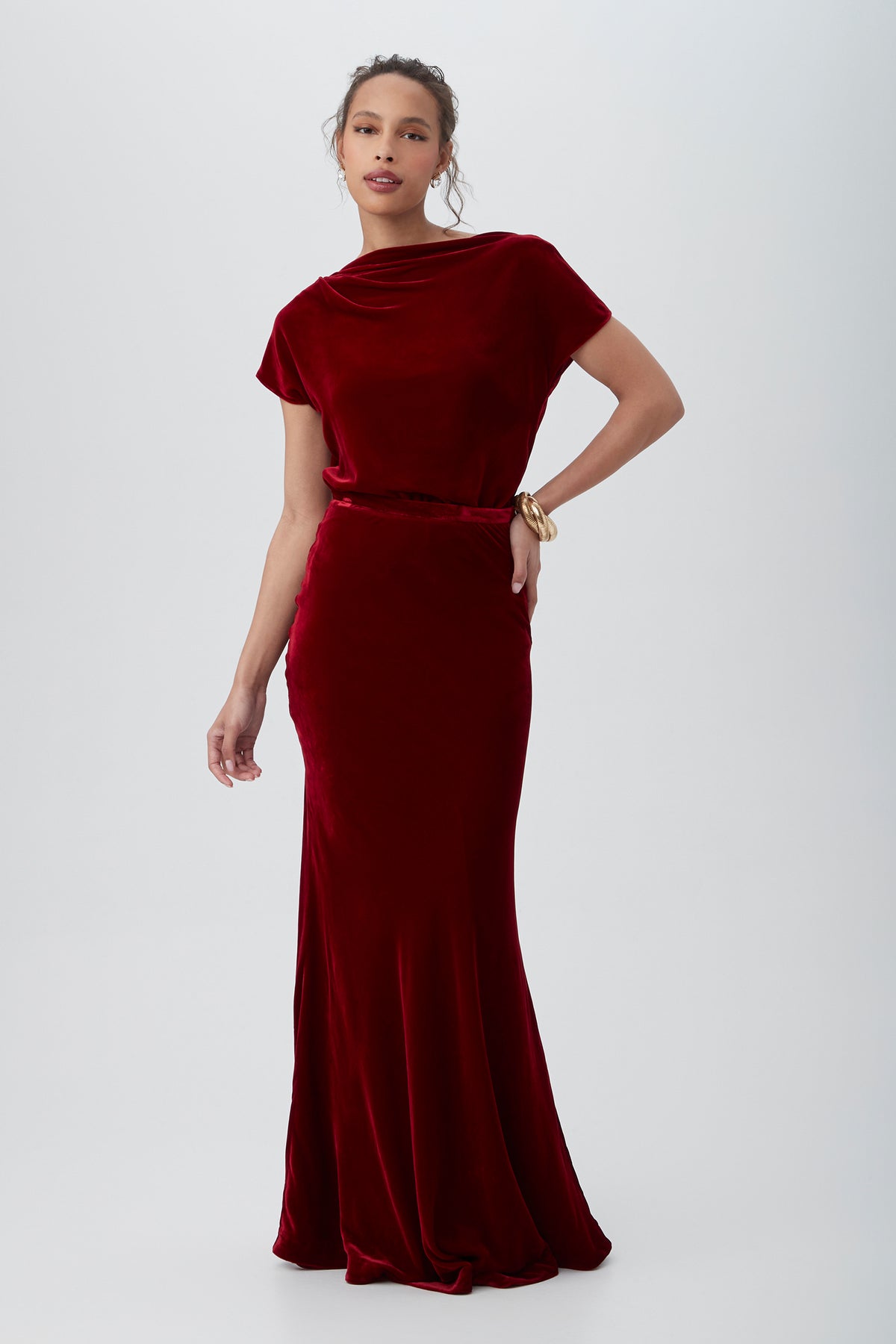 A woman stands against a plain background wearing the Rabina Velvet Mermaid Maxi Skirt in dark red, paired with a fitted top and gold bracelet, her hand on her hip showcasing the skirts elegant, floor-length silhouette.
