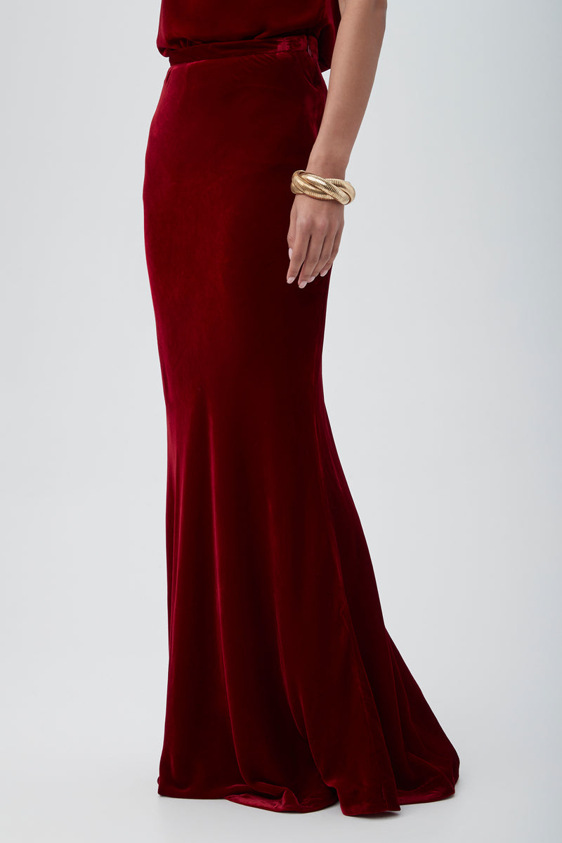 Wearing the Rabina Velvet Mermaid Maxi Skirt in deep red and a gold bracelet, a person stands against a plain light background.