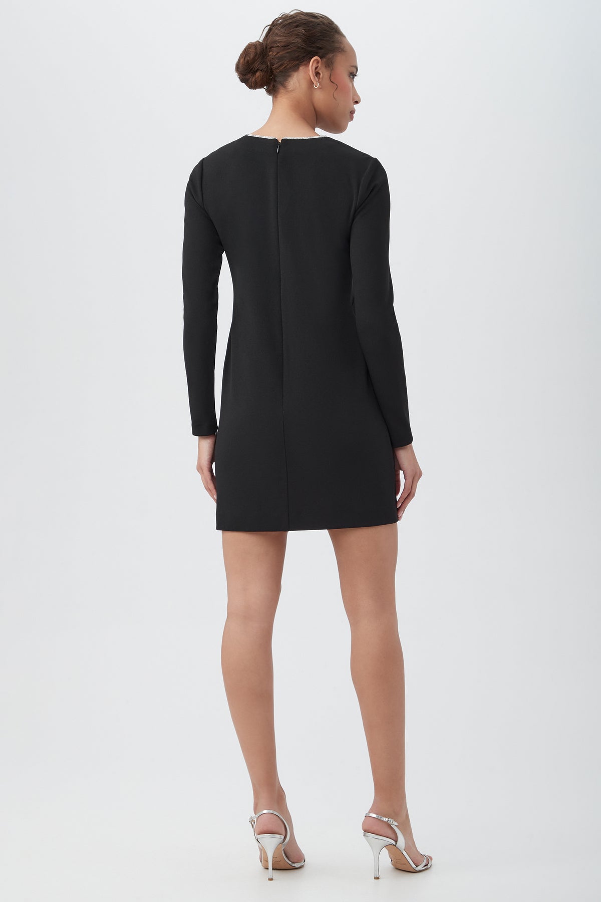 A woman with her hair in a bun faces away, wearing the Valentine Long Sleeve Sheath Dress With Rhinestone Trim and silver high-heeled sandals against a plain light background.
