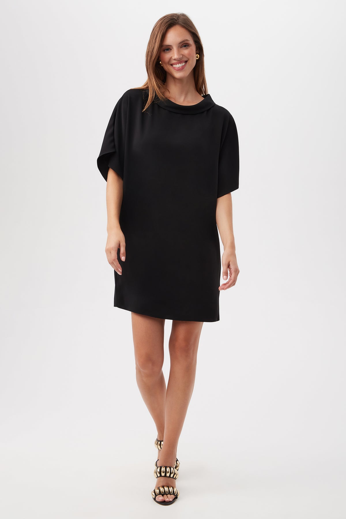 A woman smiles in the Honeysuckle Short Dolman Sleeve Shift Dress in black, featuring a loose fit and short sleeves. She pairs it with black and gold sandals, her straight brown hair falling to her shoulders against a plain white background.