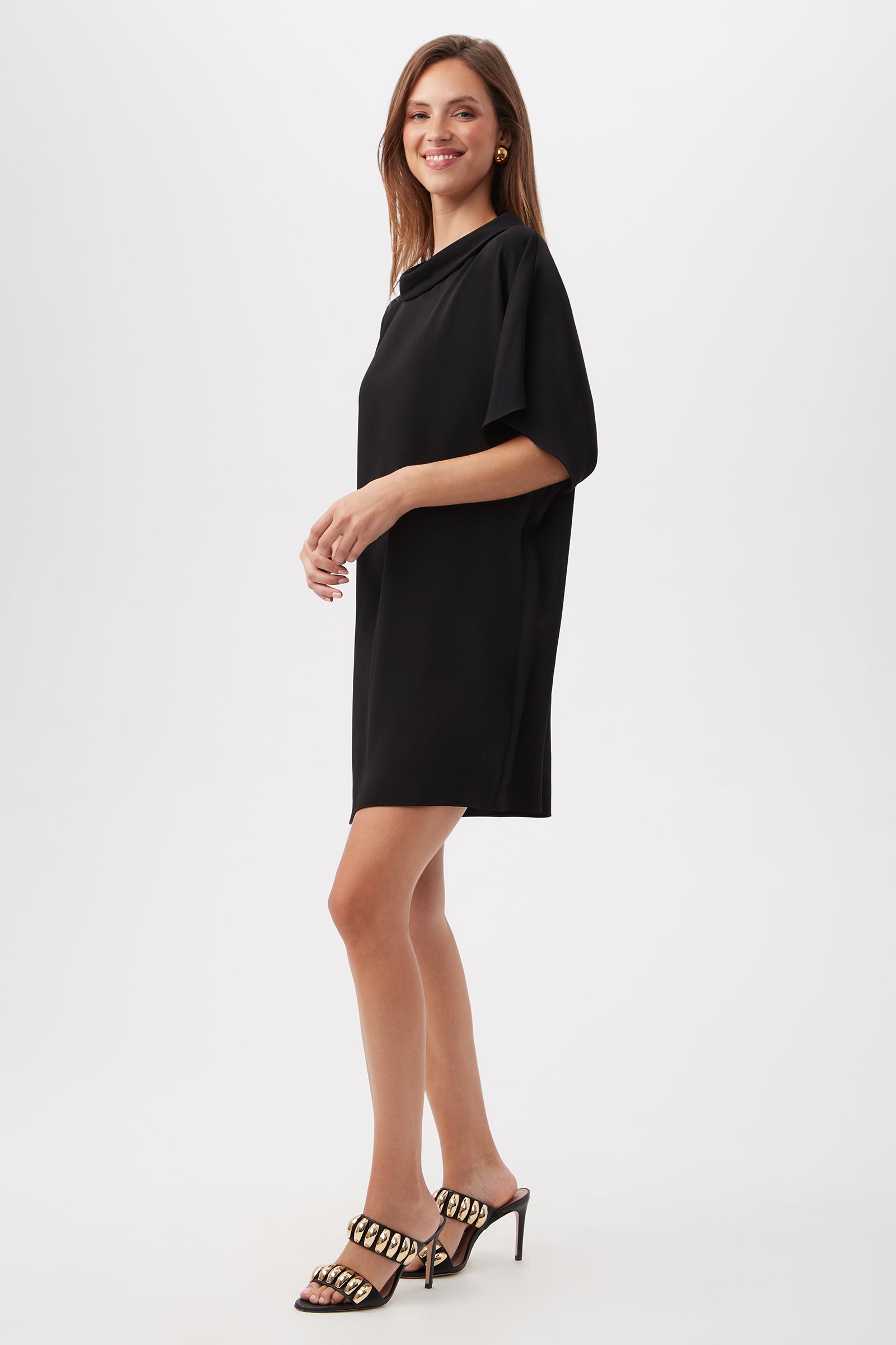 A woman smiles in the Honeysuckle Short Dolman Sleeve Shift Dress, paired with heeled sandals, against a plain white background.