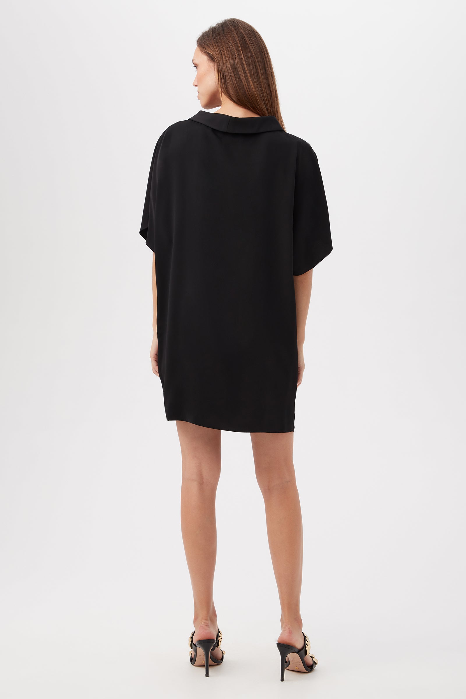 A woman with straight brown hair stands facing away, wearing the Honeysuckle Short Dolman Sleeve Shift Dress and black high-heeled sandals, set against a plain white background.