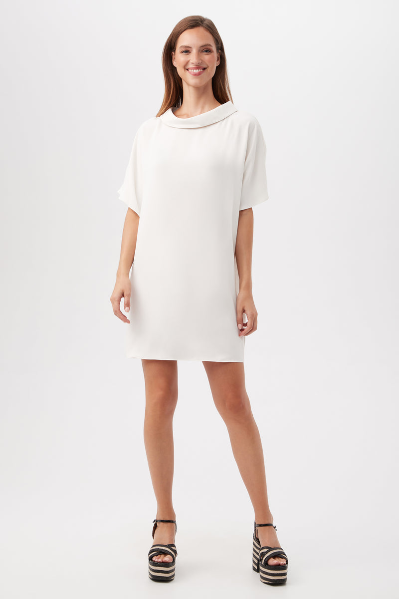 A woman smiles in the Honeysuckle Short Dolman Sleeve Shift Dress in Winter White, paired with black platform sandals, against a plain white background.