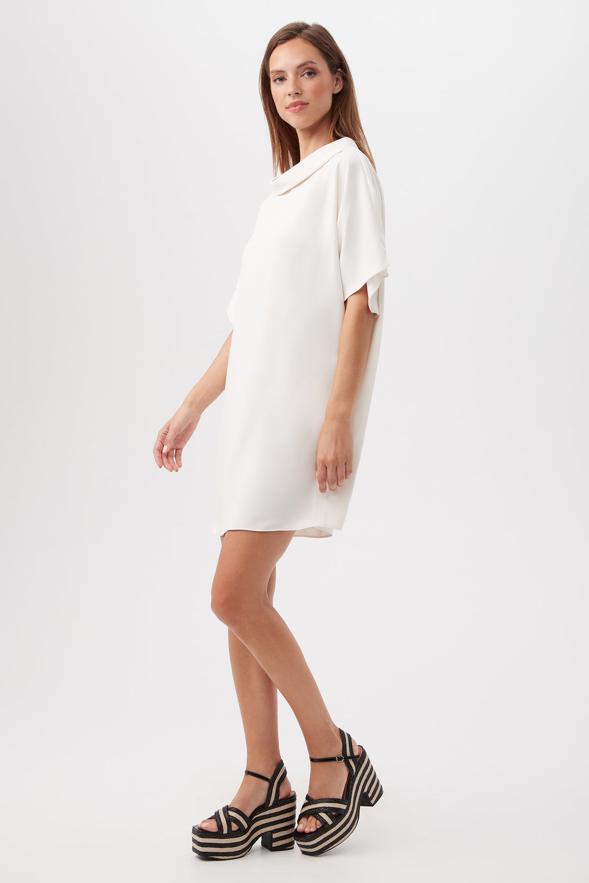 A woman stands against a white background wearing the Honeysuckle Short Dolman Sleeve Shift Dress, a loose-fitting, short-sleeved white dress, paired with black platform sandals that feature striped soles. She faces slightly to the side.