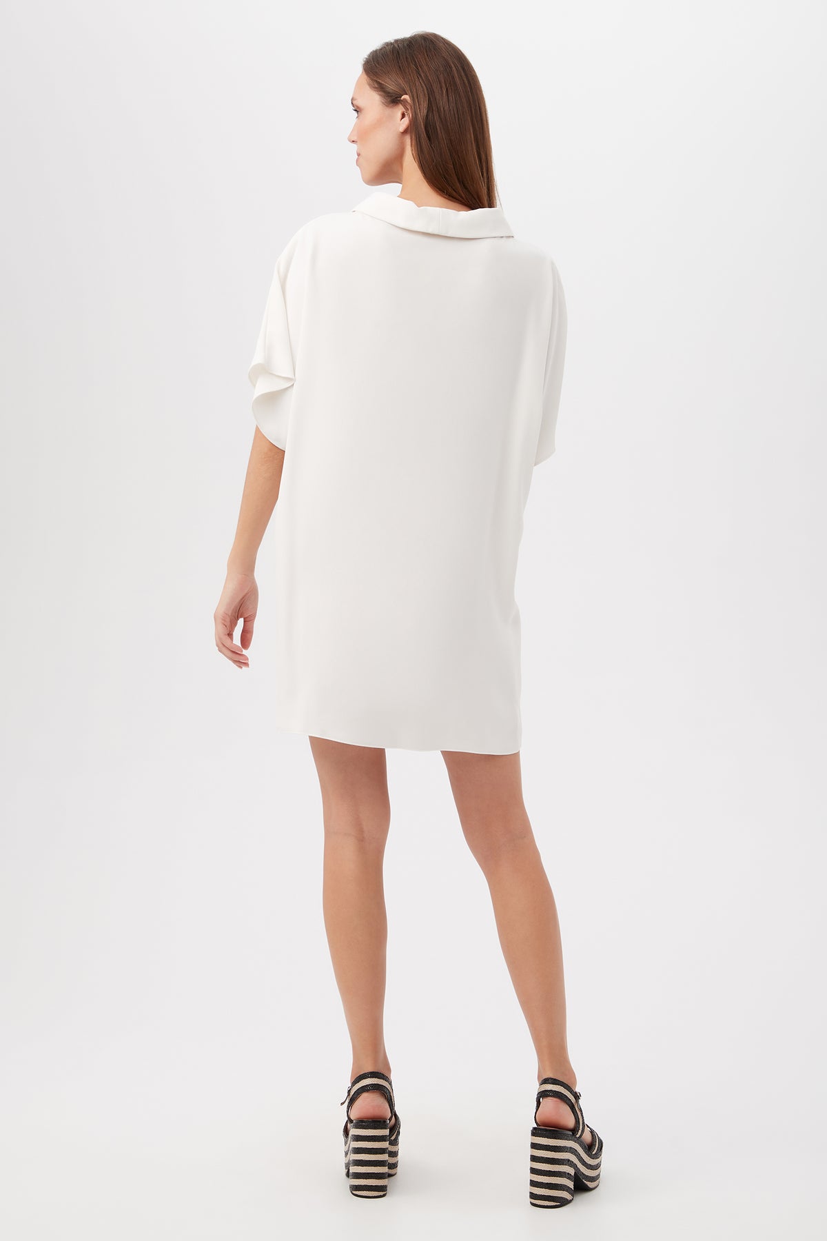 A woman faces away, wearing the Honeysuckle Short Dolman Sleeve Shift Dress in white with short sleeves, paired with black-and-white platform sandals against a plain white background.