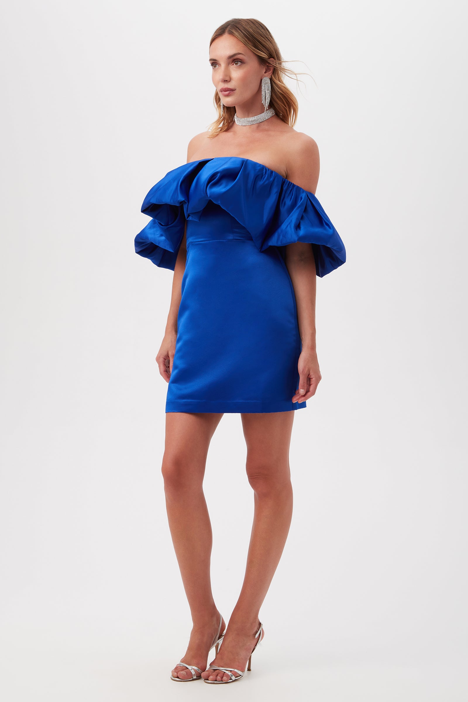 A woman stands against a white background wearing the Nia Satin Off The Shoulder Short Cocktail Dress in shiny royal blue, styled with dramatic ruffled sleeves, silver high-heeled sandals, large earrings, and a sparkling choker necklace.