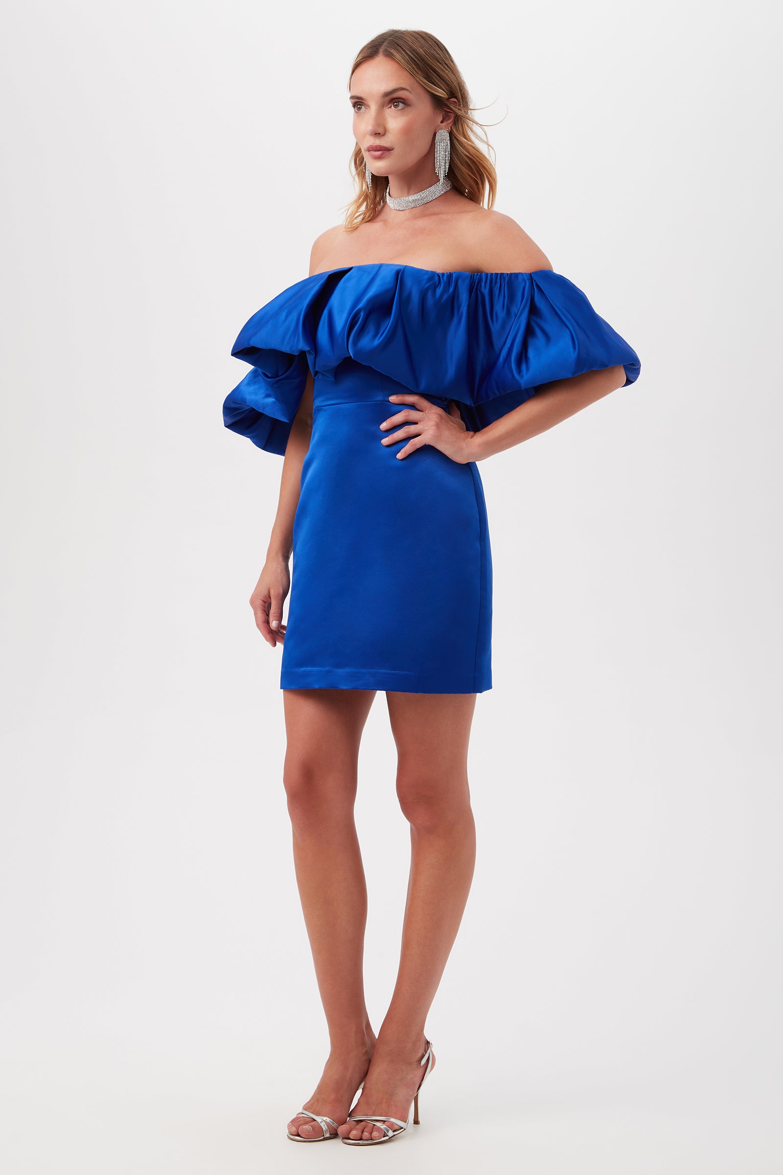 A woman models the Nia Satin Off The Shoulder Short Cocktail Dress in vibrant blue, featuring dramatic ruffled sleeves, styled with silver high-heeled sandals, bold earrings, and a statement choker against a white background.
