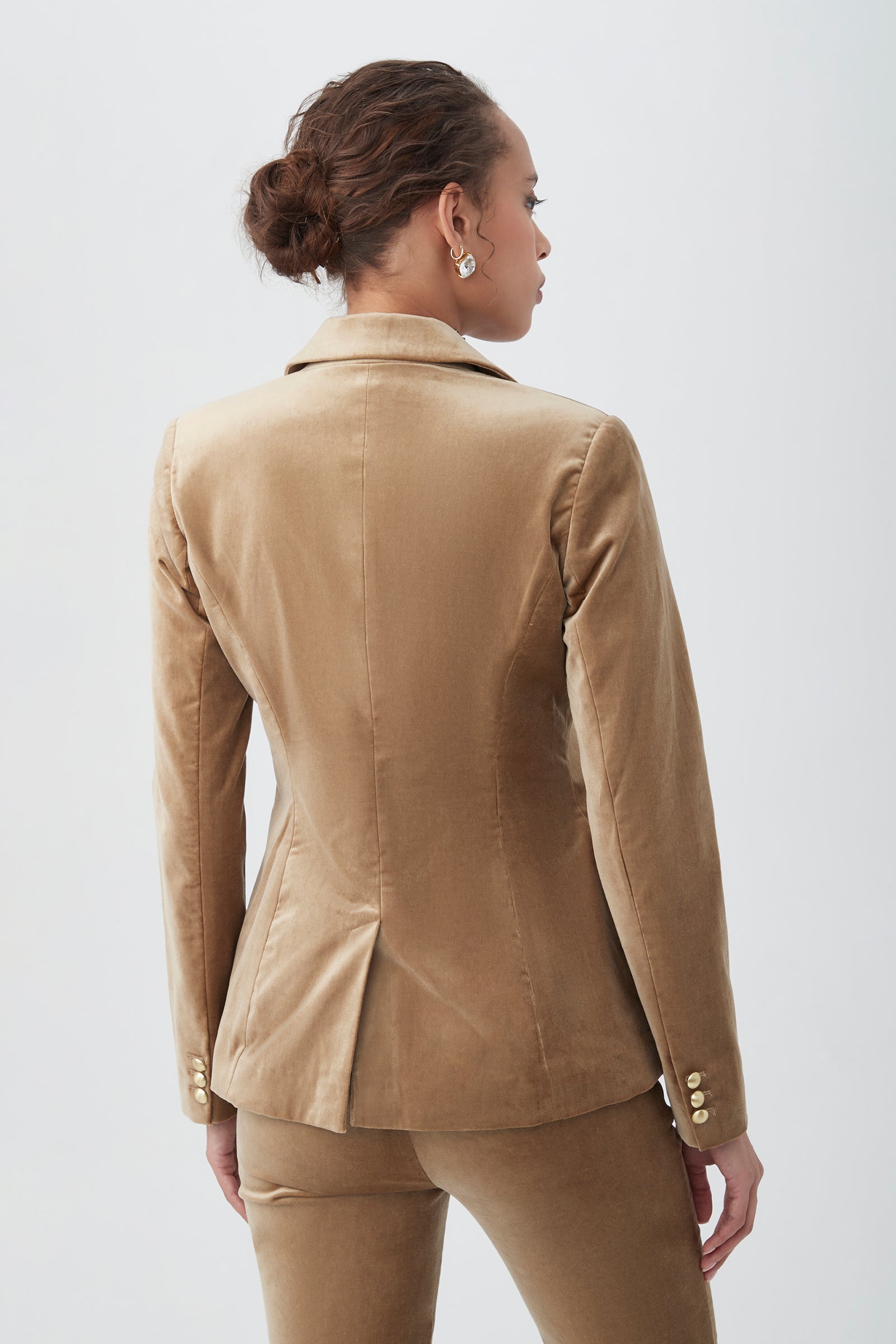 A woman with brown hair in a bun is seen from behind, wearing the Midlands Velveteen Blazer Jacket in tan and matching pants. Gold buttons accent the blazer's sleeves as she stands against a plain light background.