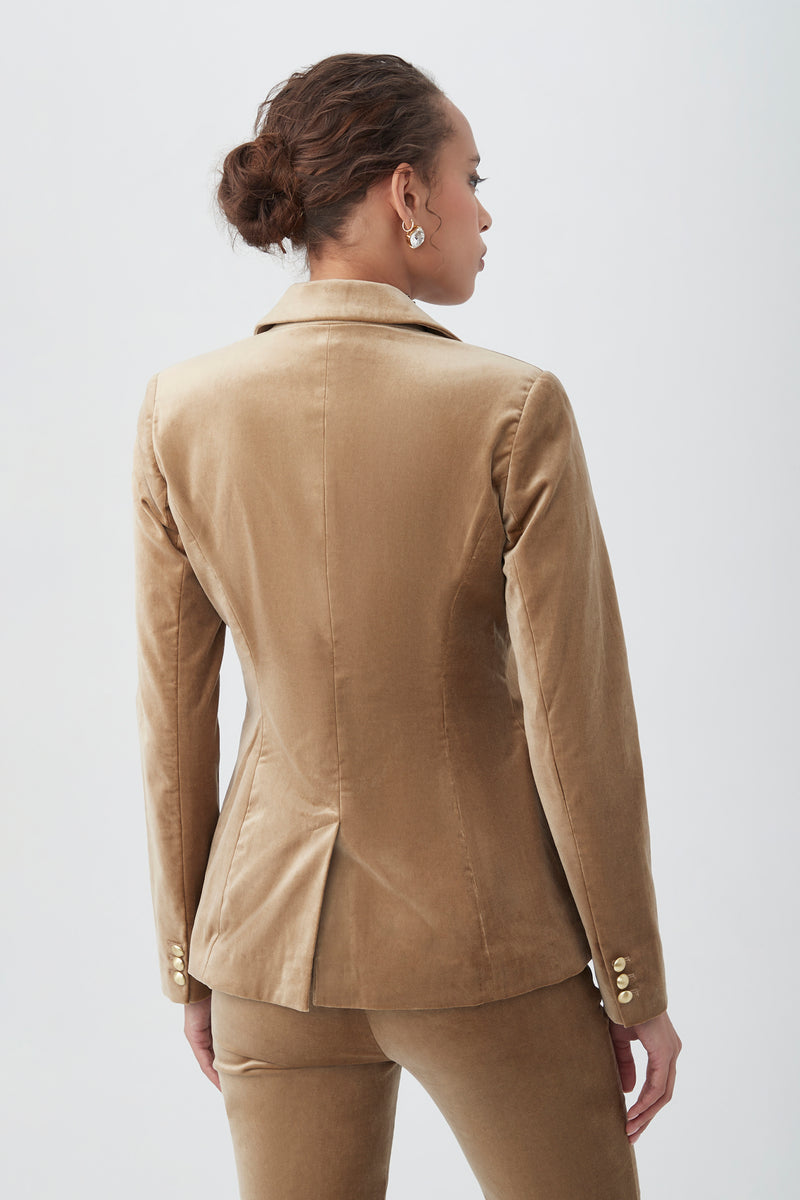 A woman with brown hair in a bun is seen from behind, wearing the Midlands Velveteen Blazer Jacket in tan and matching pants. Gold buttons accent the blazer's sleeves as she stands against a plain light background.