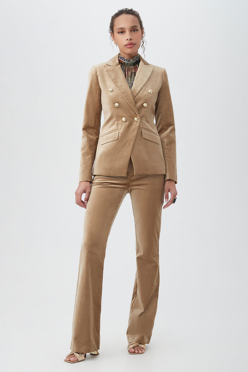 A woman stands against a plain background wearing the Midlands Velveteen Blazer Jacket in tan with gold buttons, paired with matching flared pants, a printed turtleneck, and open-toed heels.