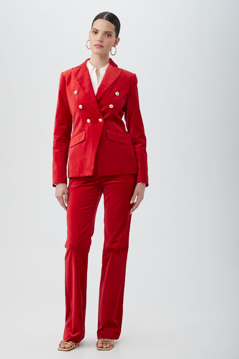 A woman stands against a plain background wearing the Midlands Velveteen Blazer Jacket in red, styled with gold buttons, a white blouse, hoop earrings, open-toed heels, and her hair pulled back with a neutral expression.