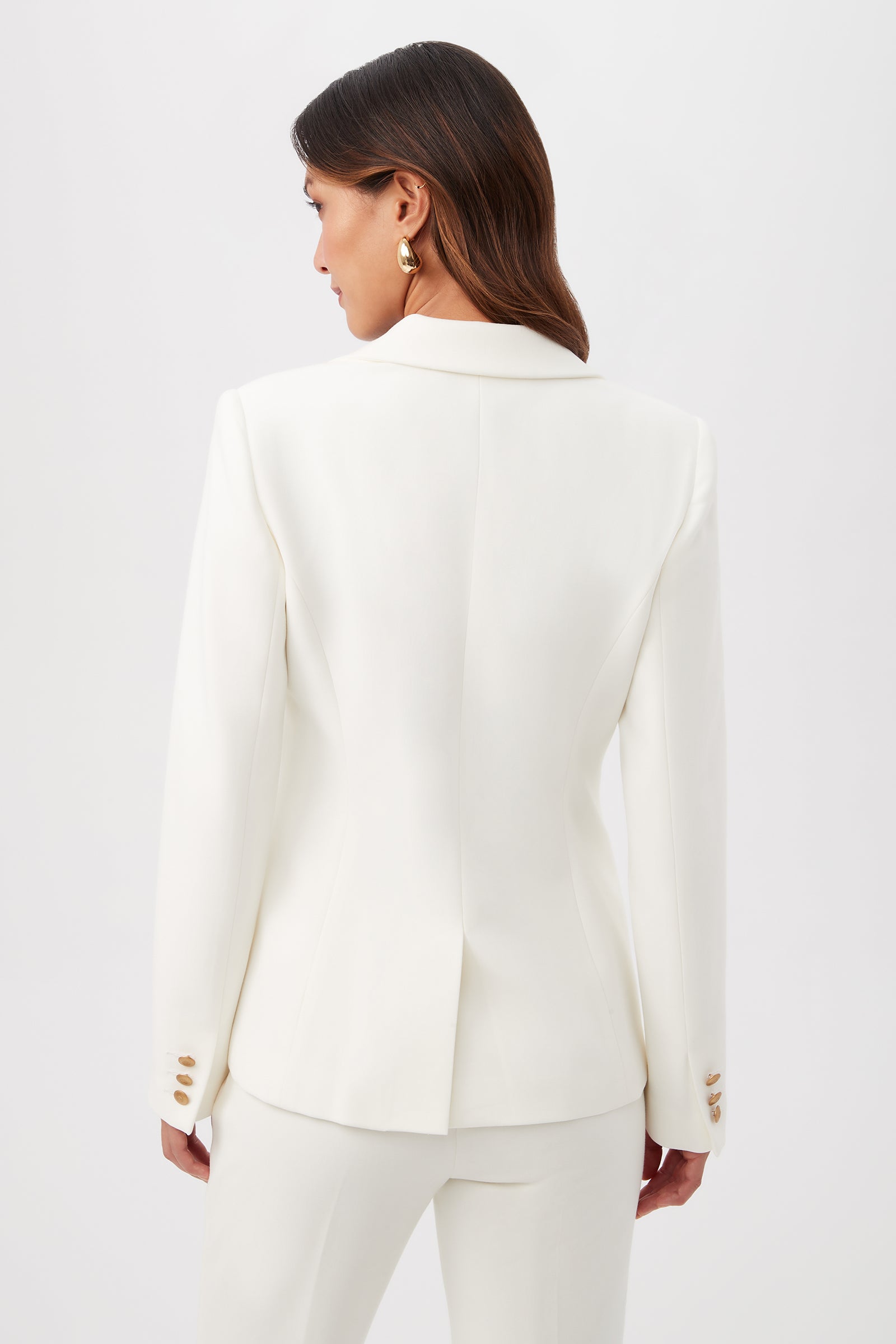 A woman with long brown hair, wearing gold earrings and the Midlands Luxe Drape Stretch Blazer Jacket with matching white pants, stands facing away from the camera against a plain white background.