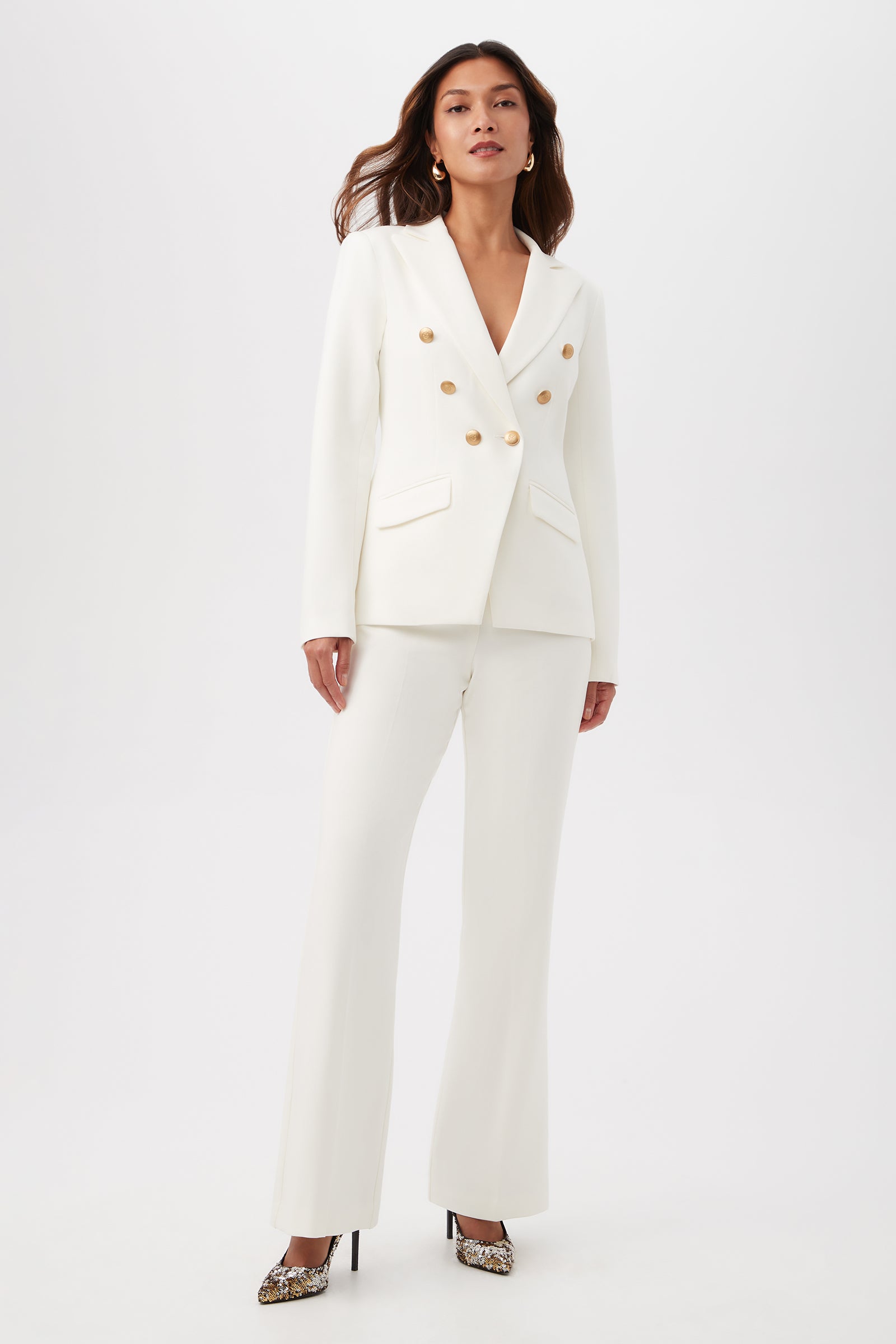 A woman stands confidently in WINTER WHITE Lancelot 2 Luxe Drape Bootcut Pants, styled with gold accents and snakeskin high heels, against a plain white background.