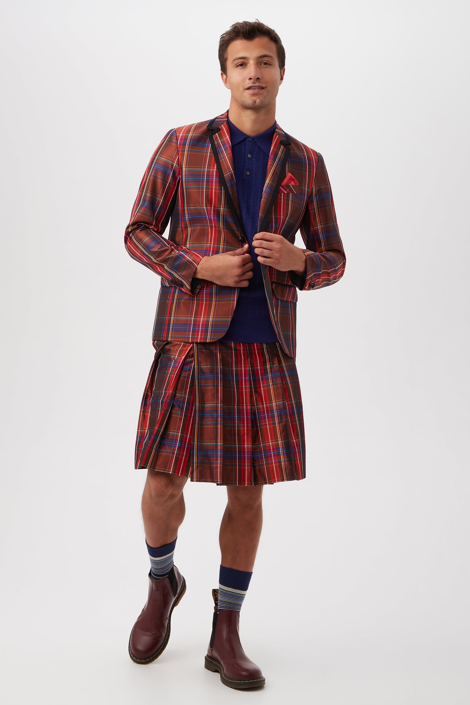 A man stands against a plain background wearing the Livingston Pleated Kilt in plaid taffeta with a matching blazer, navy polo shirt, striped socks, and brown boots.