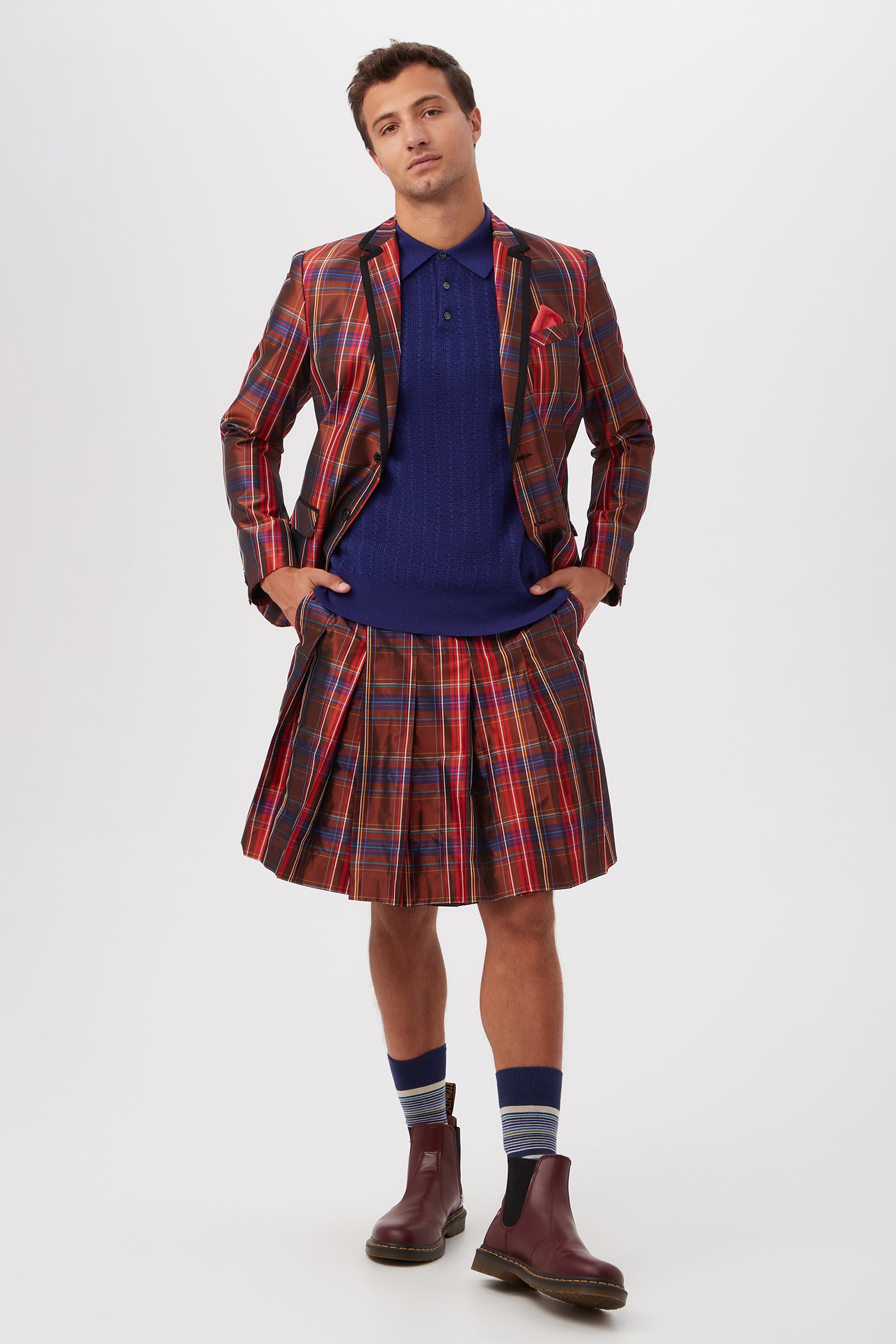 A man stands against a white background wearing the Livingston Pleated Kilt - Plaid Taffeta, a matching red plaid blazer, blue knit shirt, striped blue socks, and burgundy boots. His hands are in his pockets with a neutral expression.