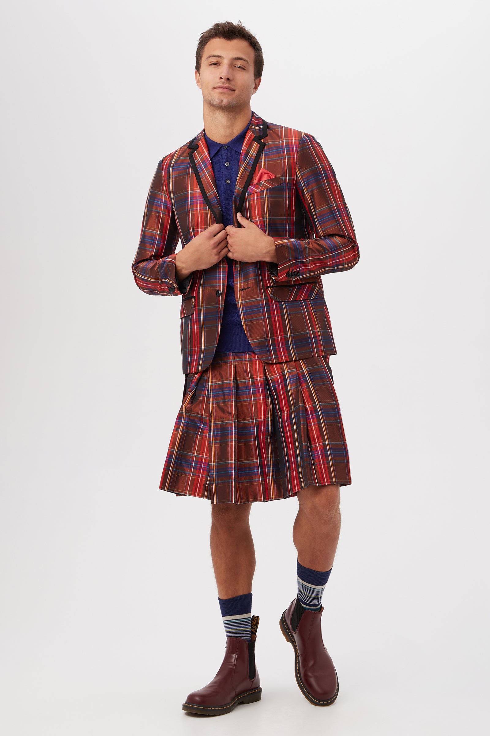 A man stands against a white background wearing the Livingston Pleated Kilt - Plaid Taffeta with a matching jacket, dark blue shirt, striped socks, and brown ankle boots. He holds his jacket lapels and looks confidently at the camera.