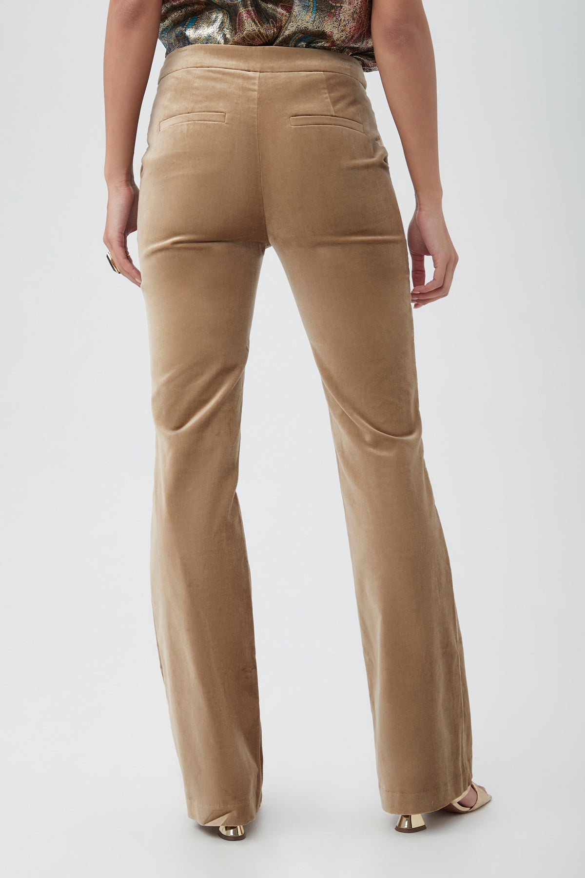 A person stands facing away from the camera, wearing the Westport Velveteen Bootcut Pant—high-waisted tan velvet pants with two back pockets and flared legs. Their arms are at their sides, paired with open-toed shoes.
