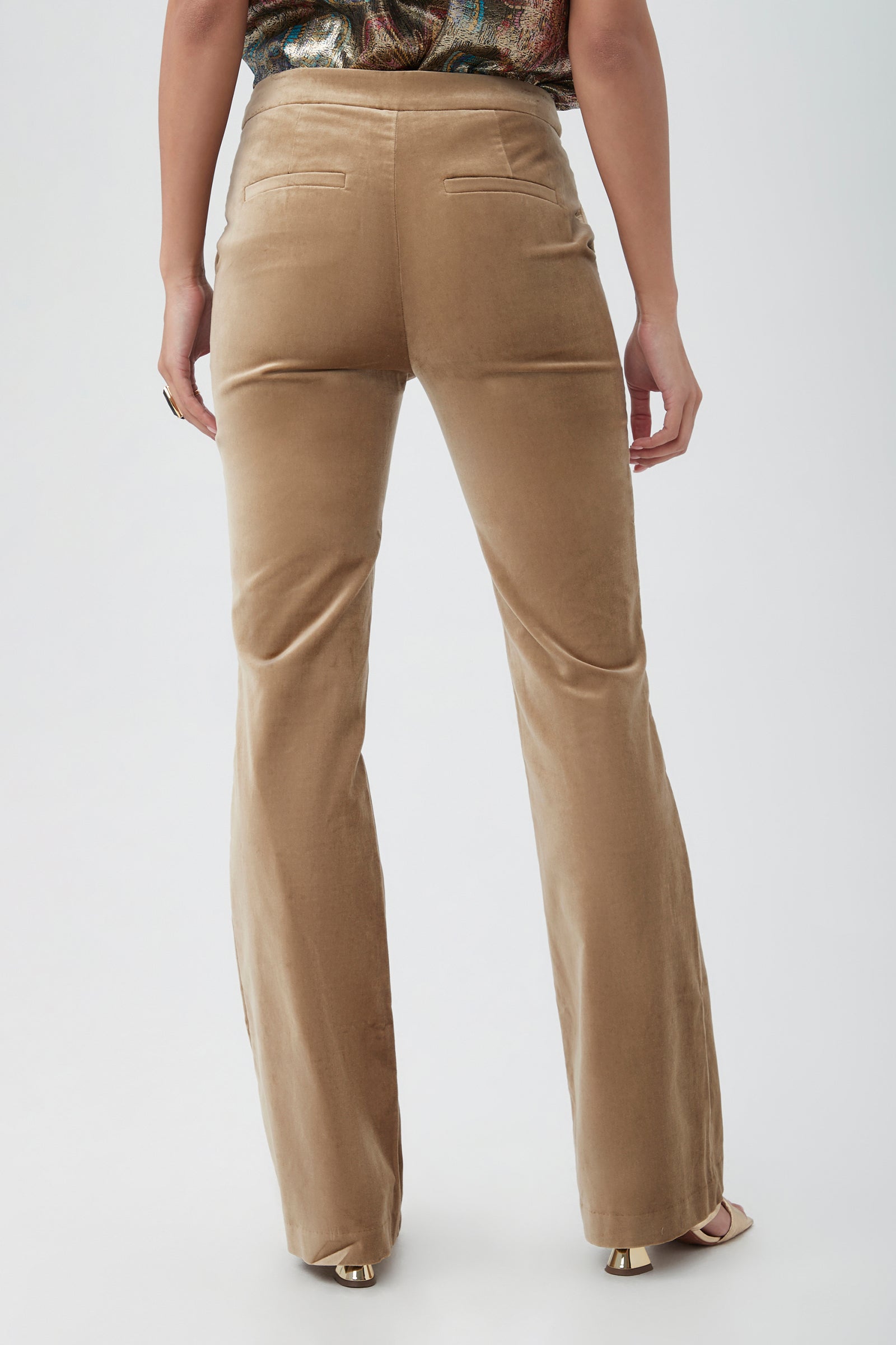 A person stands facing away from the camera, wearing the Westport Velveteen Bootcut Pant—high-waisted tan velvet pants with two back pockets and flared legs. Their arms are at their sides, paired with open-toed shoes.