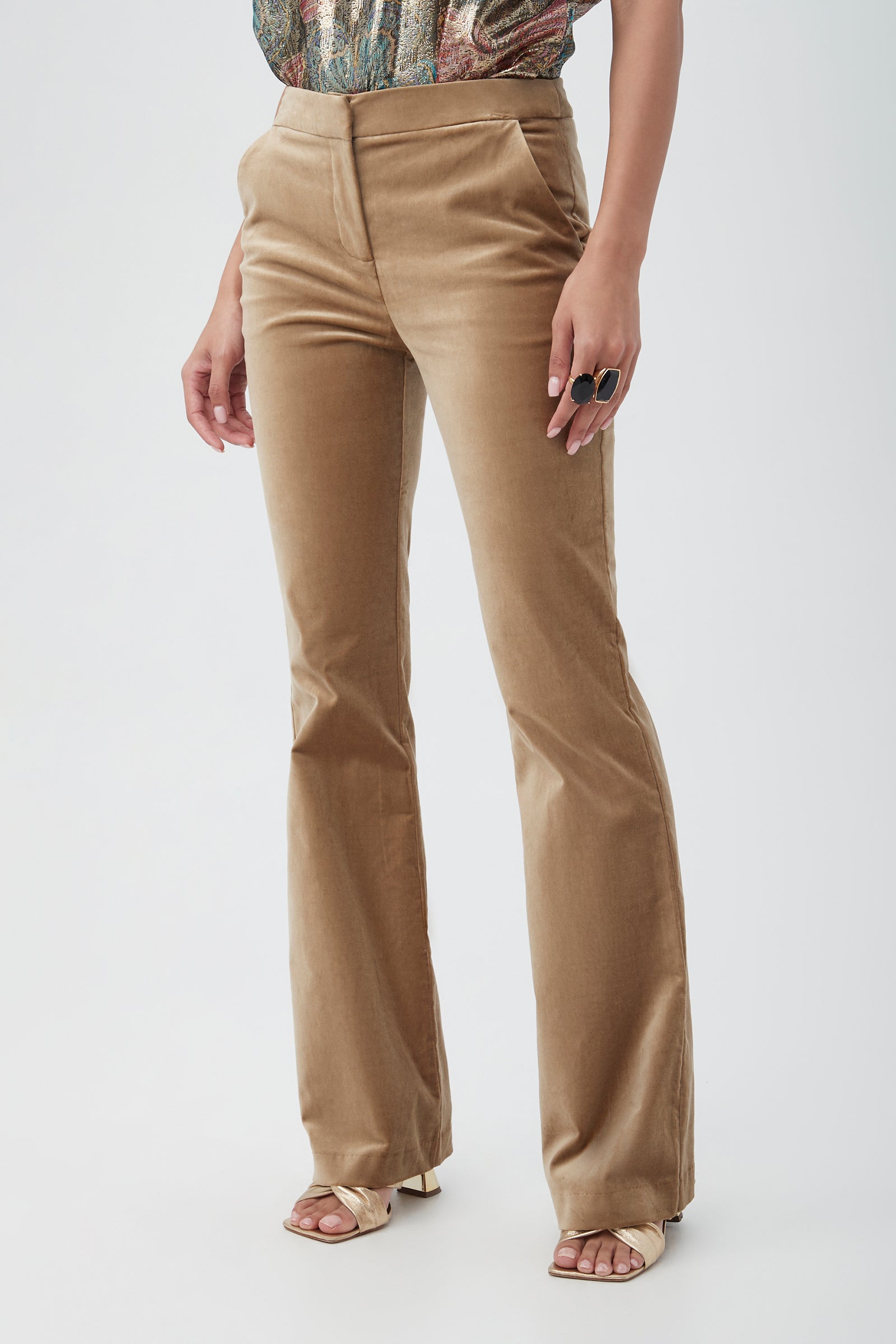 A person wearing Westport Velveteen Bootcut Pants in tan, a patterned top, open-toe heeled sandals, and a large black ring stands against a plain light background.