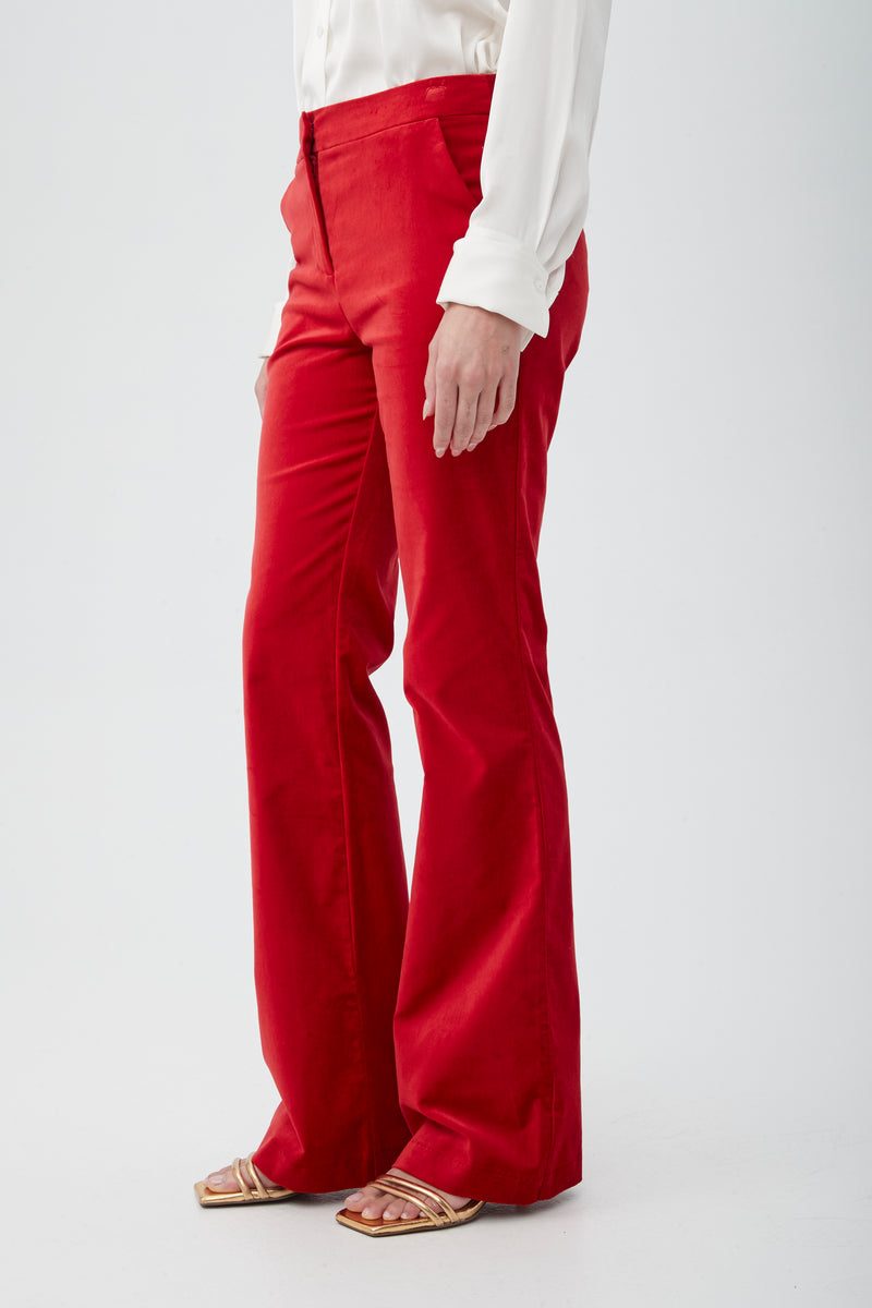 A person models the Westport Velveteen Bootcut Pant in bright red, paired with a white long-sleeve blouse and gold open-toe sandals against a plain white background. Only their lower body is visible.