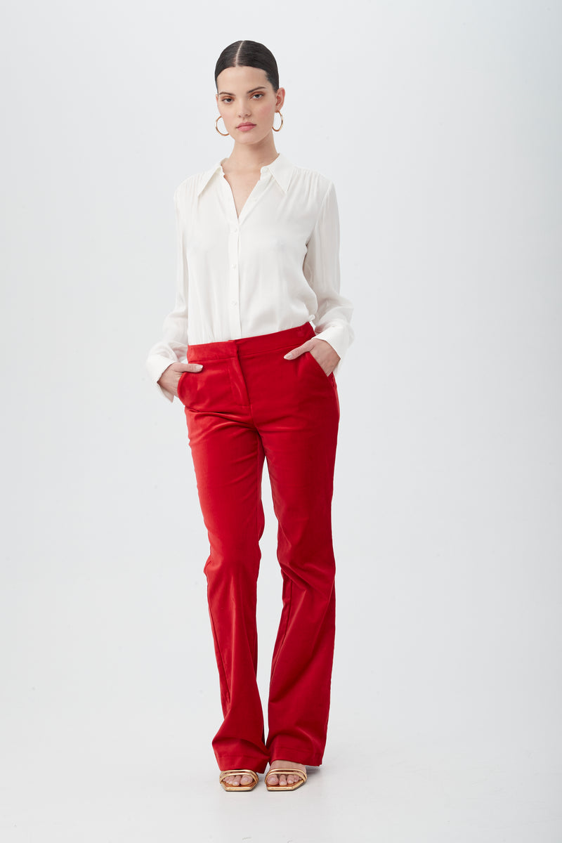 A woman stands against a plain white background, wearing the Westport Velveteen Bootcut Pant in red with a white blouse and sandals. Her dark hair is pulled back, hands in her pockets, and she looks straight ahead with a neutral expression.