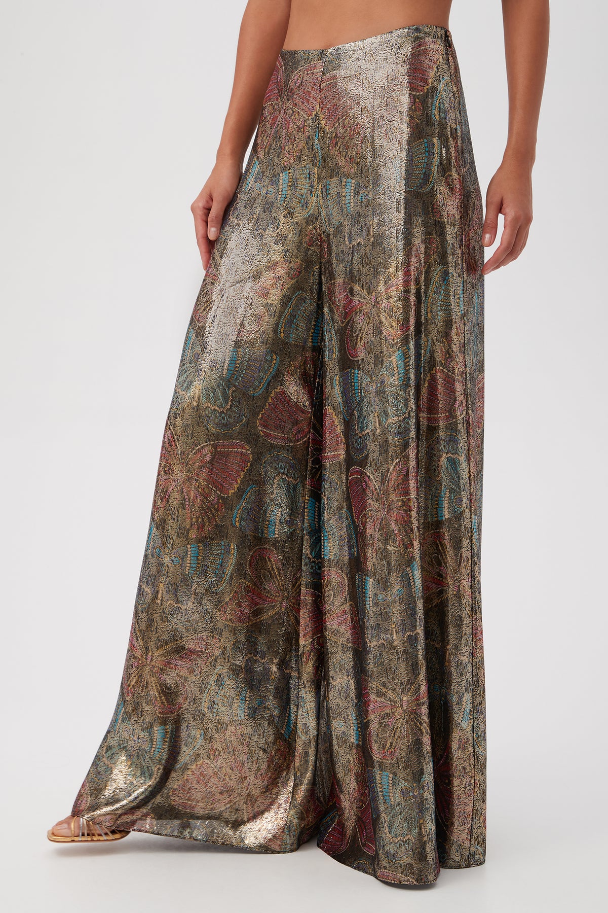 A person wearing the Siya Butterfly Print Silk Wide Leg Pant, featuring a metallic sheen with multicolored floral and abstract patterns, stands against a plain light background.