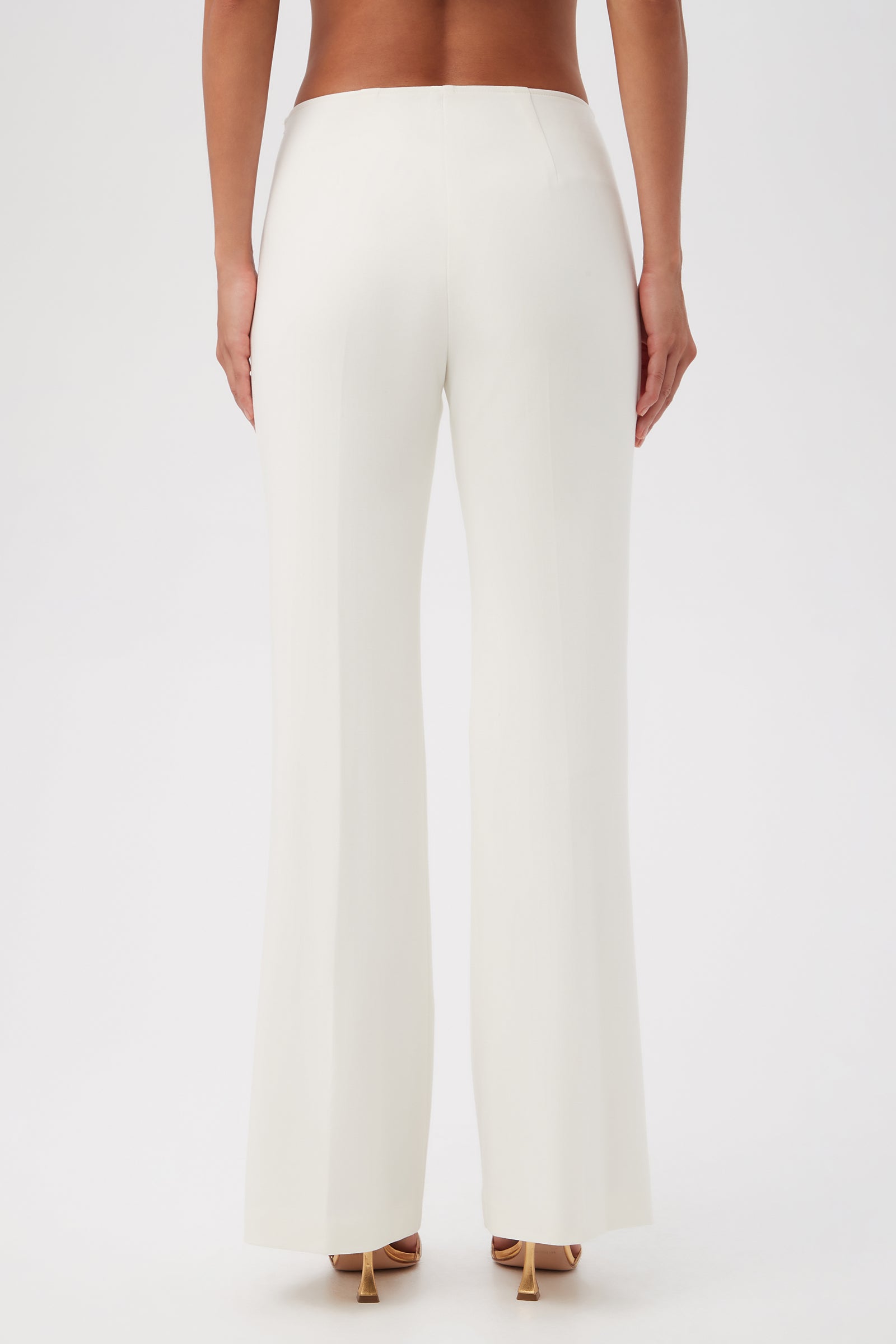 A person is shown from the back, wearing Lancelot 2 Luxe Drape Bootcut Pants in white with nude high-heeled sandals against a plain white background.