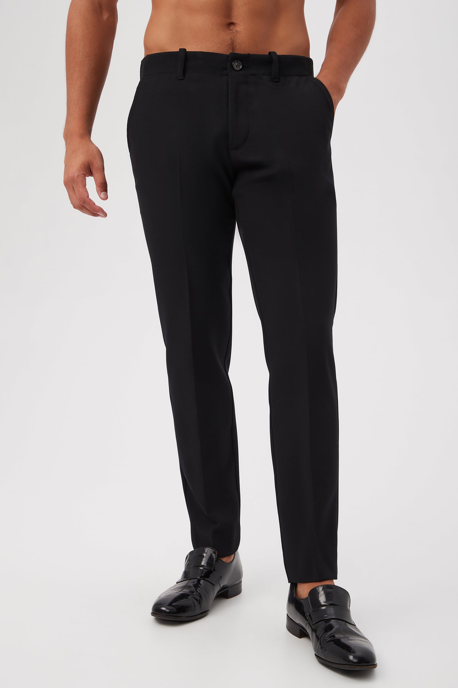 A man wearing the Alex Slim Trouser - Luxe Drape in BLACK, with his hands in the pockets and black shiny dress shoes, stands against a plain light background.