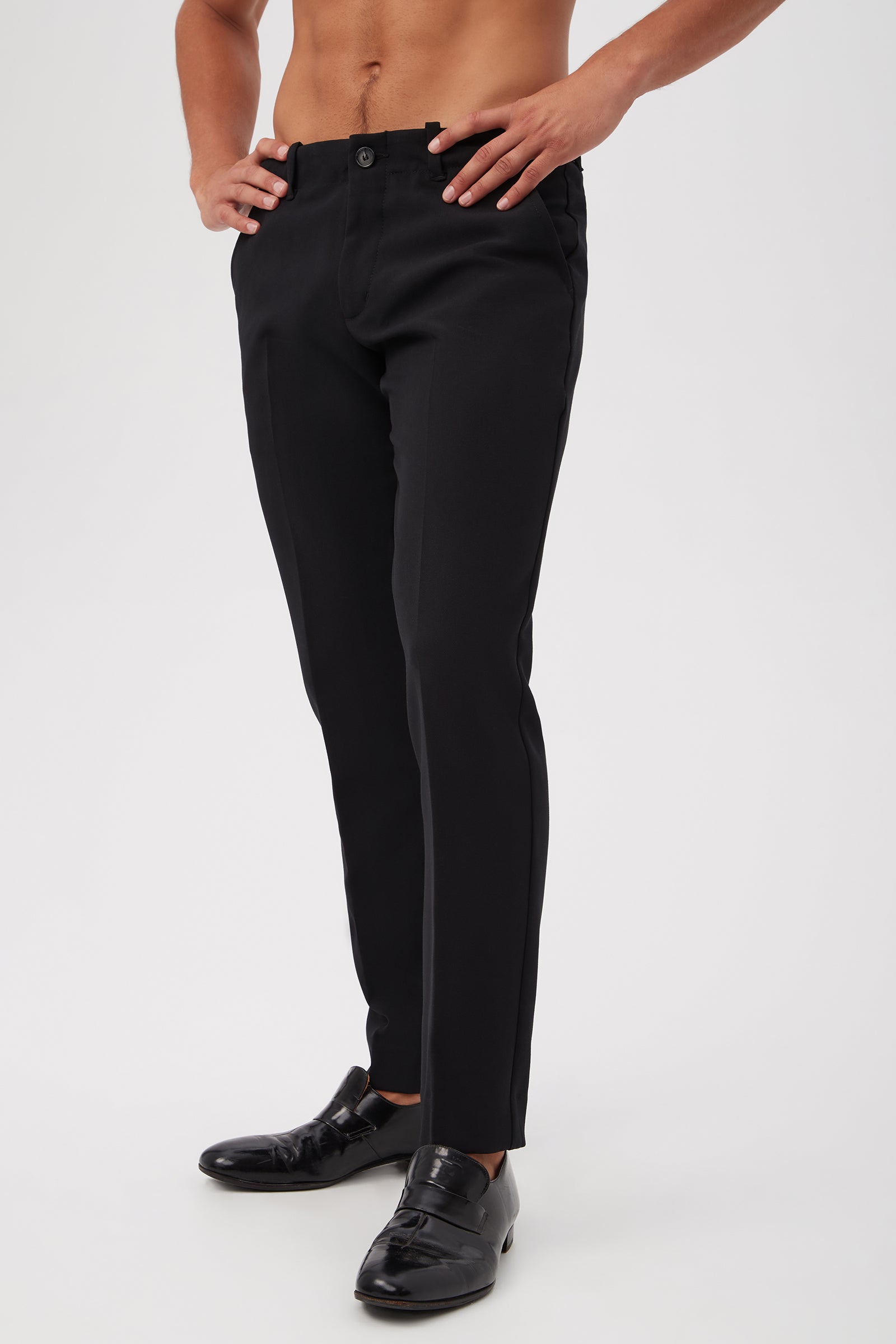 A shirtless person wearing the Alex Slim Trouser - Luxe Drape in black with shiny black loafers stands with hands on hips against a plain white background.
