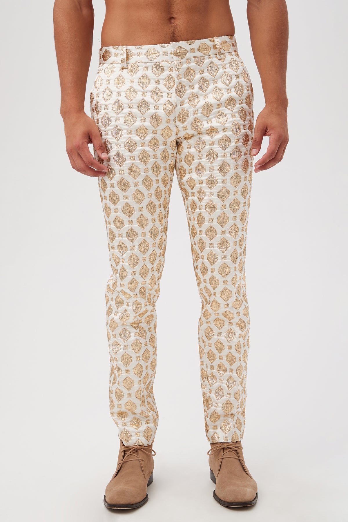 A person wears the Clyde Slim Trouser - Jubilant Jacquard, featuring shiny white fabric with a gold geometric pattern and beige dress shoes, standing against a plain white background with a bare torso.