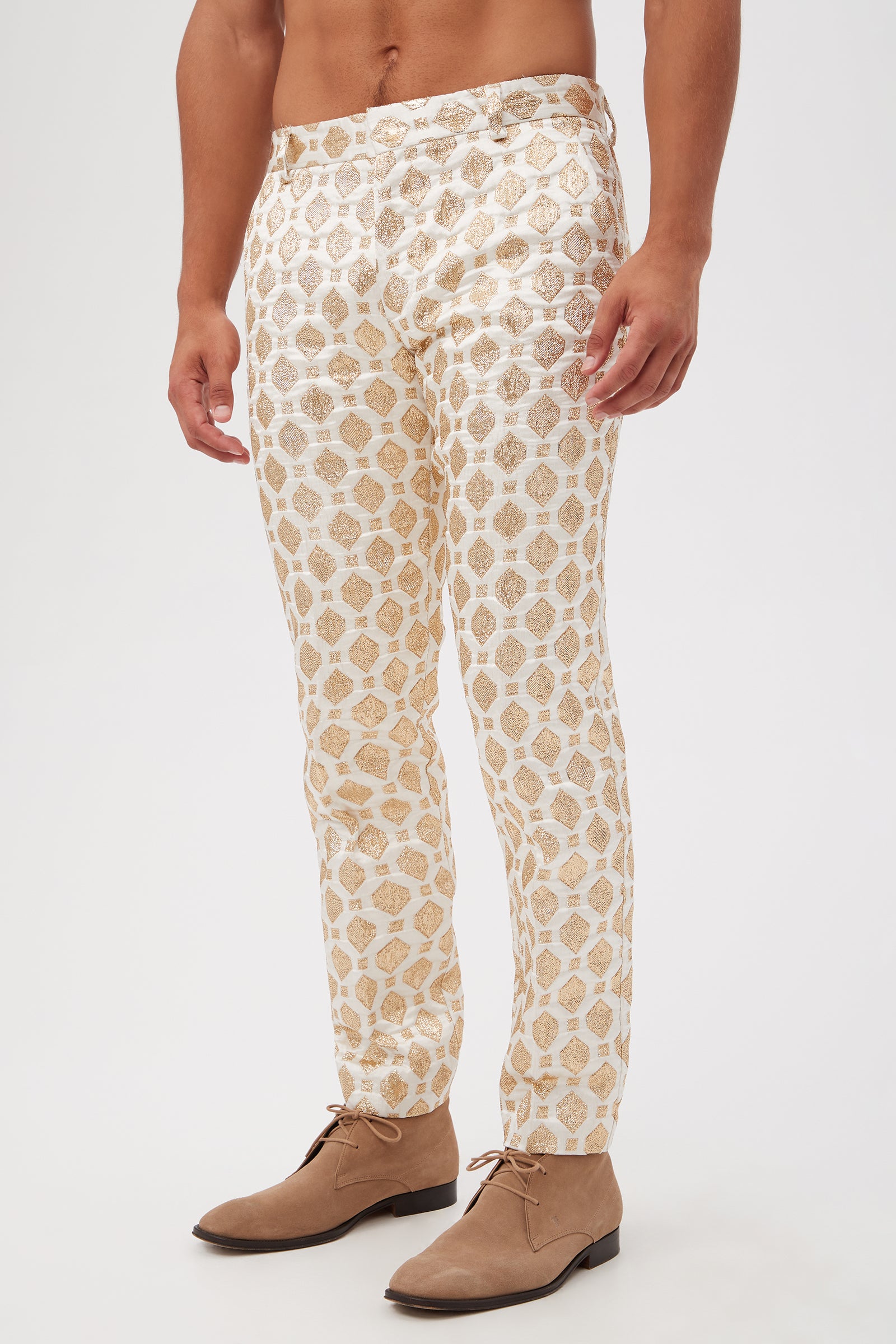 A person wears the Clyde Slim Trouser - Jubilant Jacquard, featuring a gold geometric pattern on white, paired with light brown suede lace-up shoes and standing against a plain white background. The upper body is not visible.
