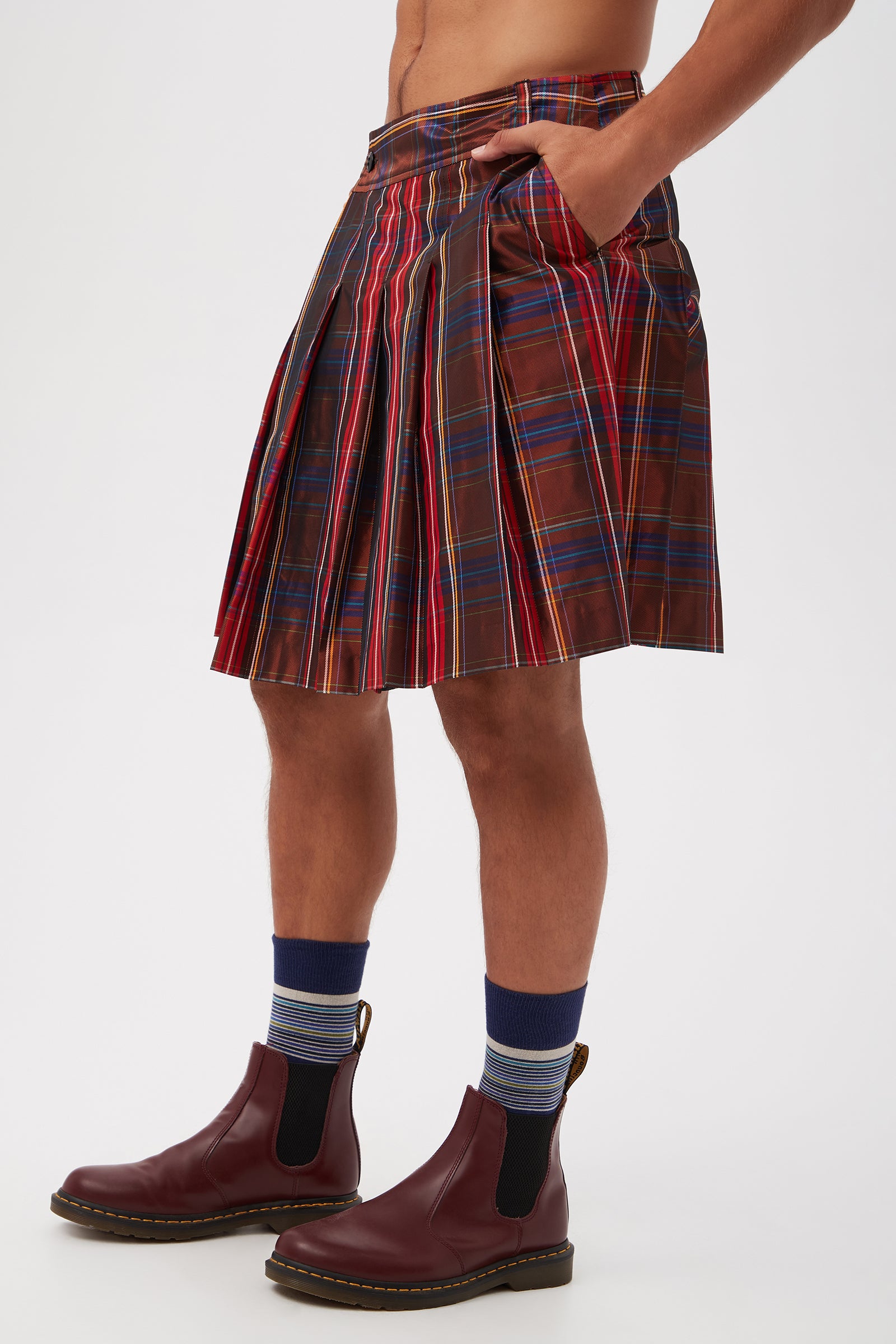 Wearing the Livingston Pleated Kilt in plaid taffeta, blue striped socks, and burgundy Chelsea boots, a shirtless person stands with hands in pockets against a plain white background, showing only their torso and legs.