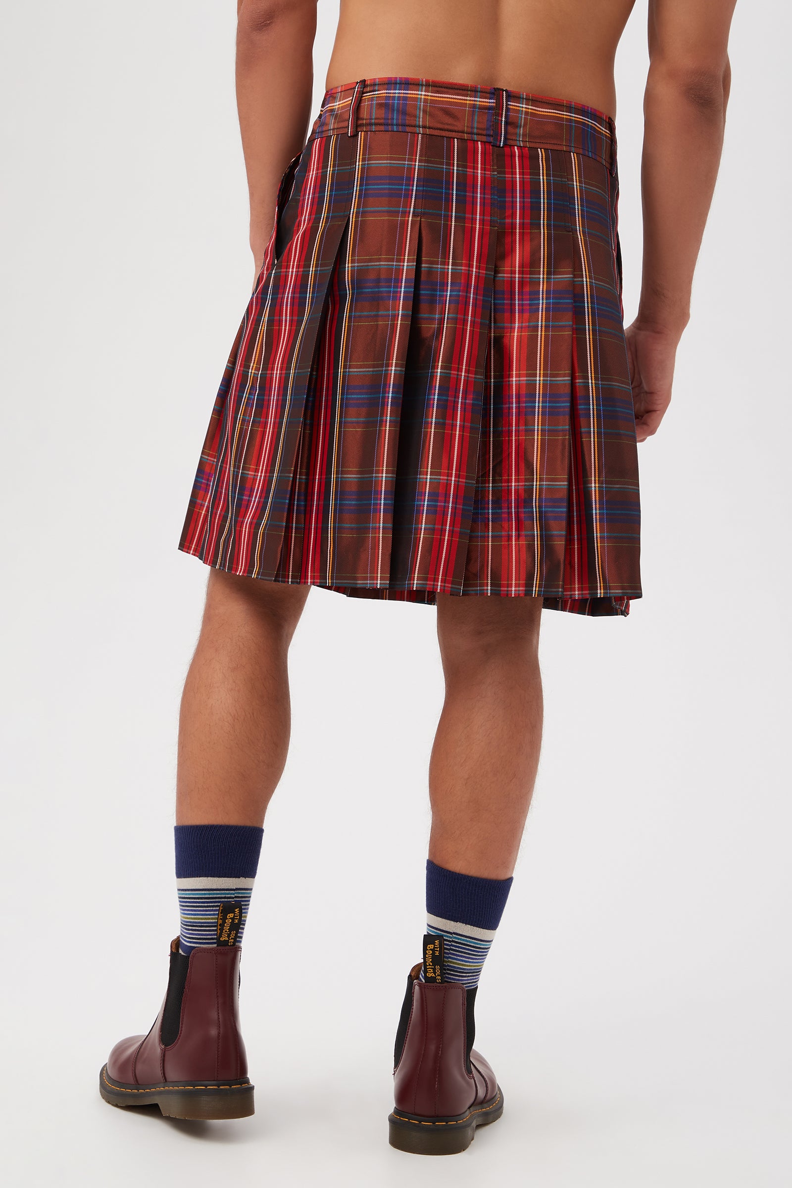A person stands with their back to the camera, wearing the Livingston Pleated Kilt - Plaid Taffeta in red and blue, paired with blue striped socks and brown boots. Their upper body is bare against a plain white background.