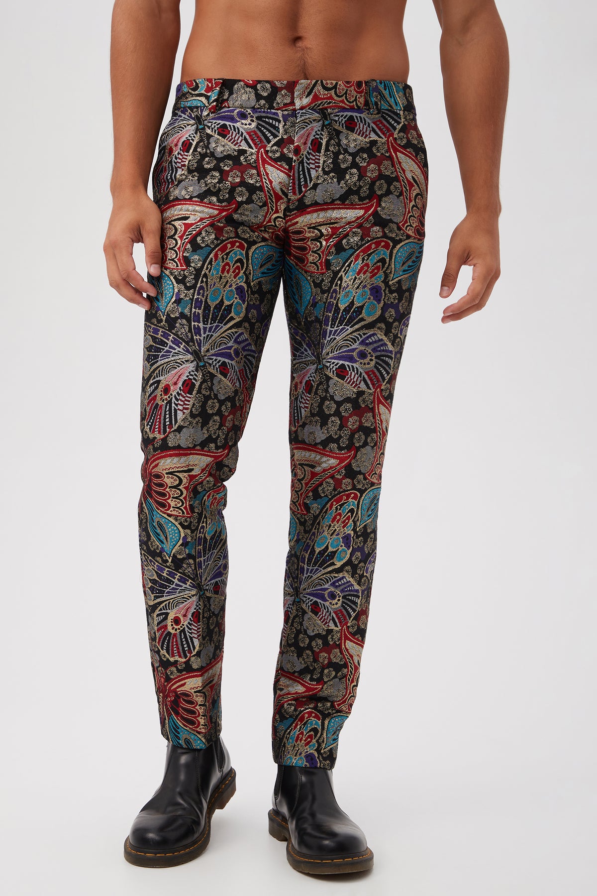 A man wearing the Clyde Slim Trouser in Monarch Jacquard, featuring a vibrant butterfly and floral pattern, paired with black Chelsea boots, stands against a plain light background. Only his lower body up to the chest is visible.