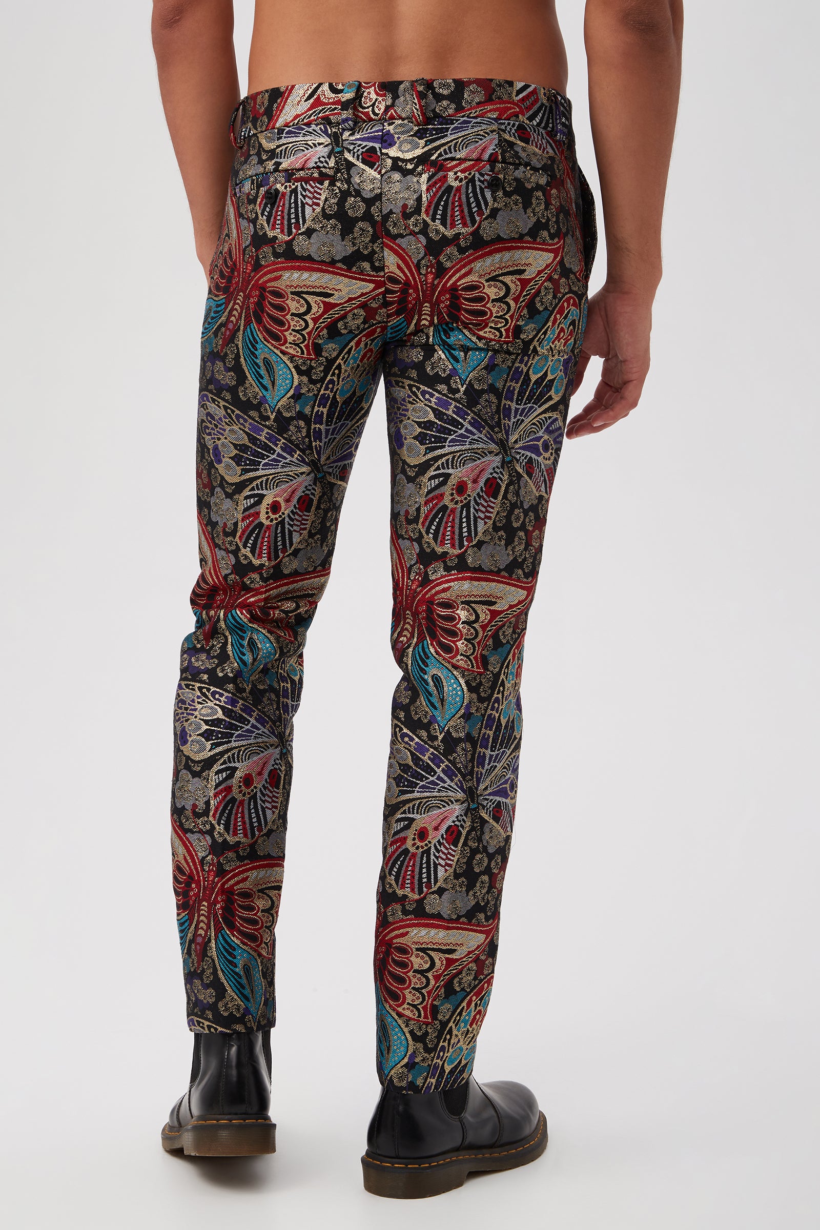 A man, shirtless and in black shoes, stands with his back to the camera wearing the Clyde Slim Trouser in Monarch Jacquard—a vibrant, multicolored butterfly and floral patterned slim-fit trouser—set against a plain white background.