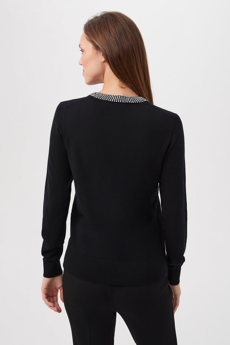 A woman with long brown hair, wearing the Laelia Merino Wool Sweater With Rhinestone Trim and black pants, stands facing away against a plain white background.